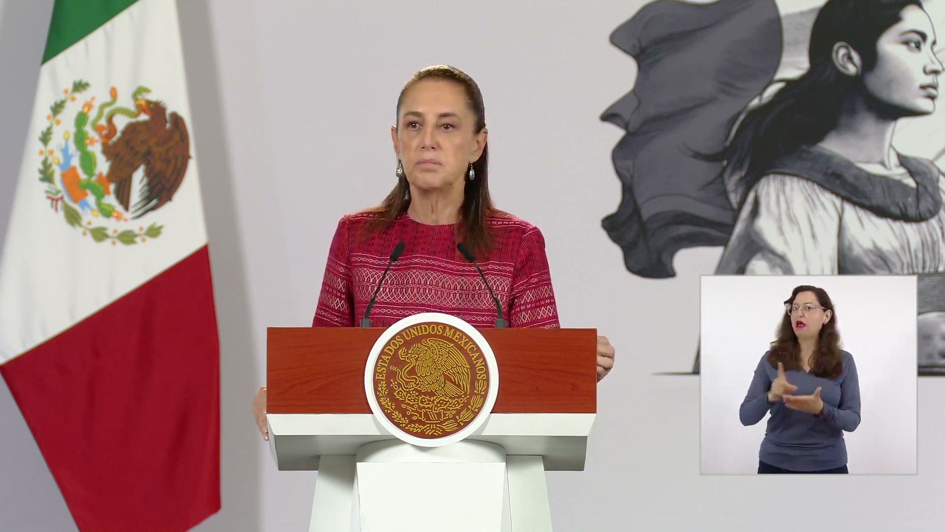 La presidenta Claudia Sheinbaum habla frente al podio con el escudo nacional, junto a una bandera de México y una intérprete de lengua de señas.