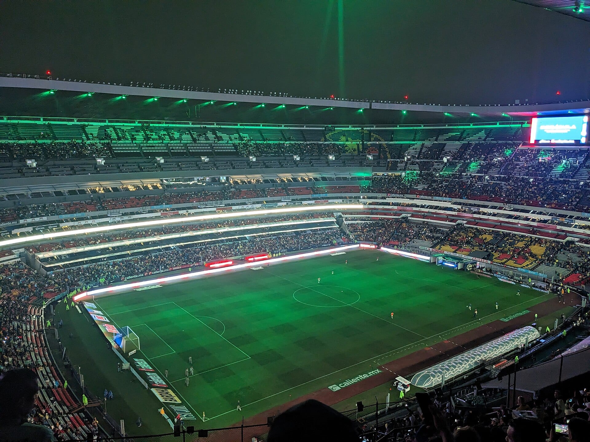 Vista nocturna del Estadio Azteca desde una grada alta, con la cancha iluminada, el techo con luces verdes y los palcos llenos de aficionados.