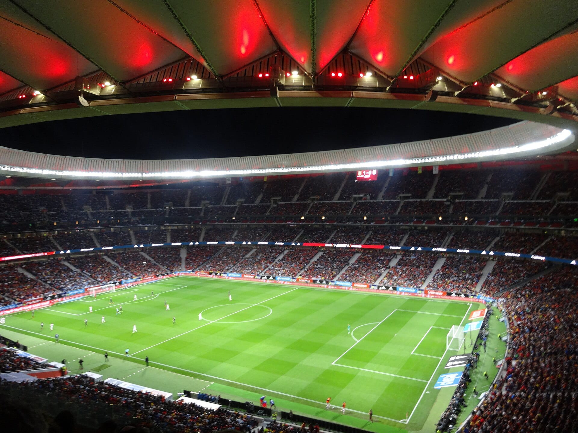 Interior de un estadio de noche, con el techo iluminado en rojo y las tribunas llenas alrededor de un campo de futbol.