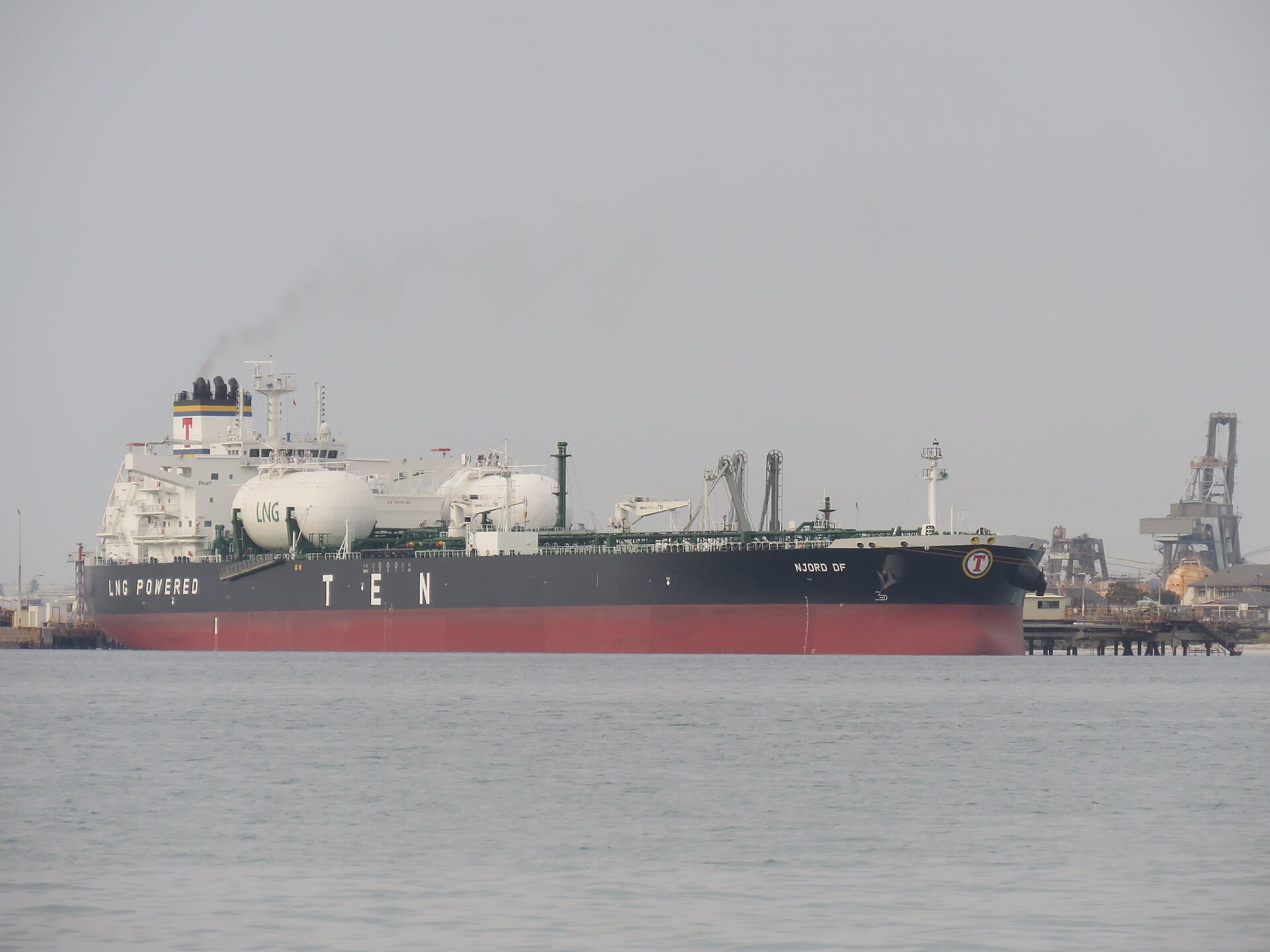 Un buque petrolero de casco negro y rojo atracado en un muelle de refinería, con grúas al fondo y el mar en calma.