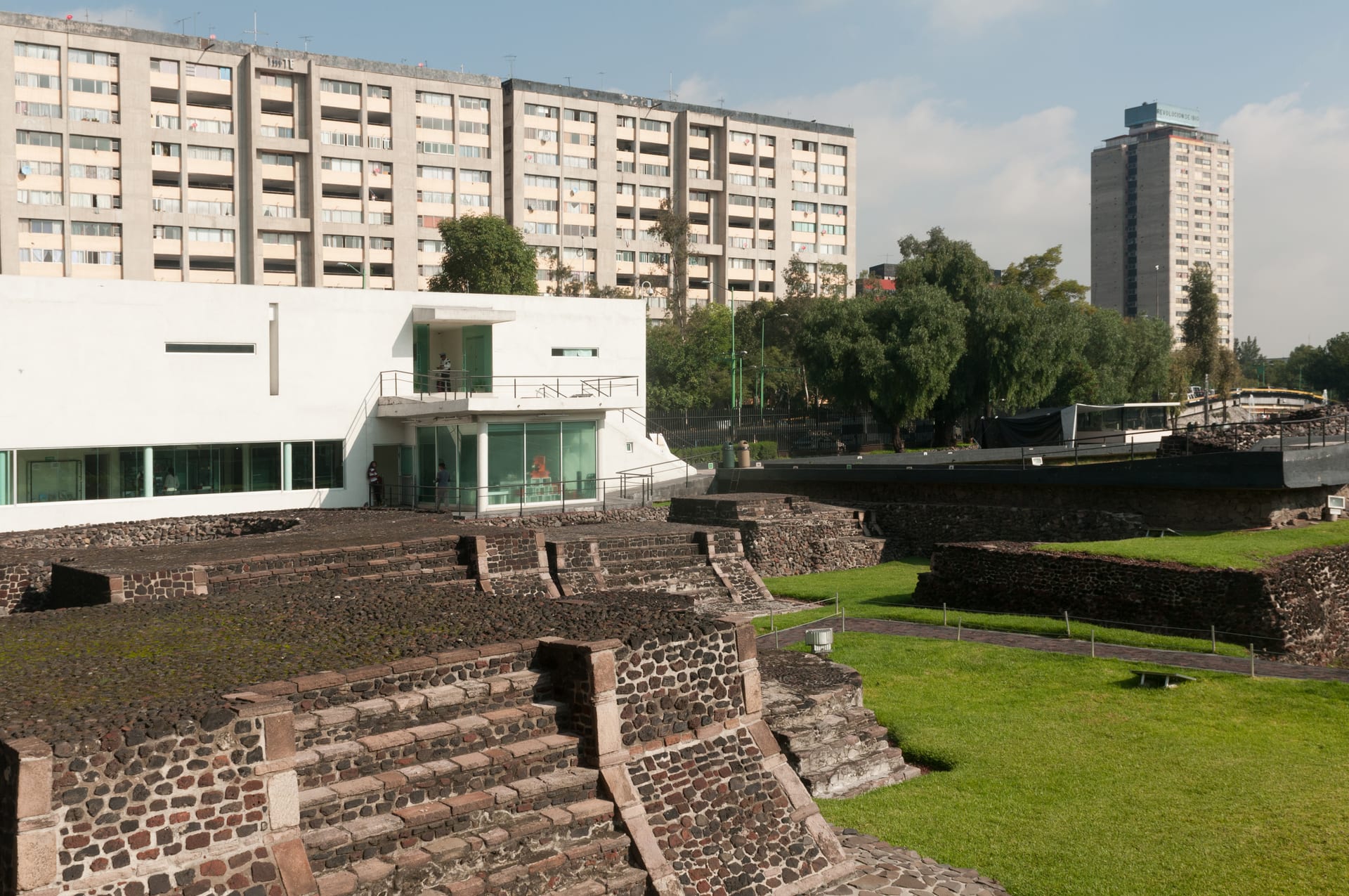 Basamentos de piedra de la zona arqueológica de Tlatelolco frente a edificios de departamentos y una torre alta, bajo un cielo parcialmente nublado.