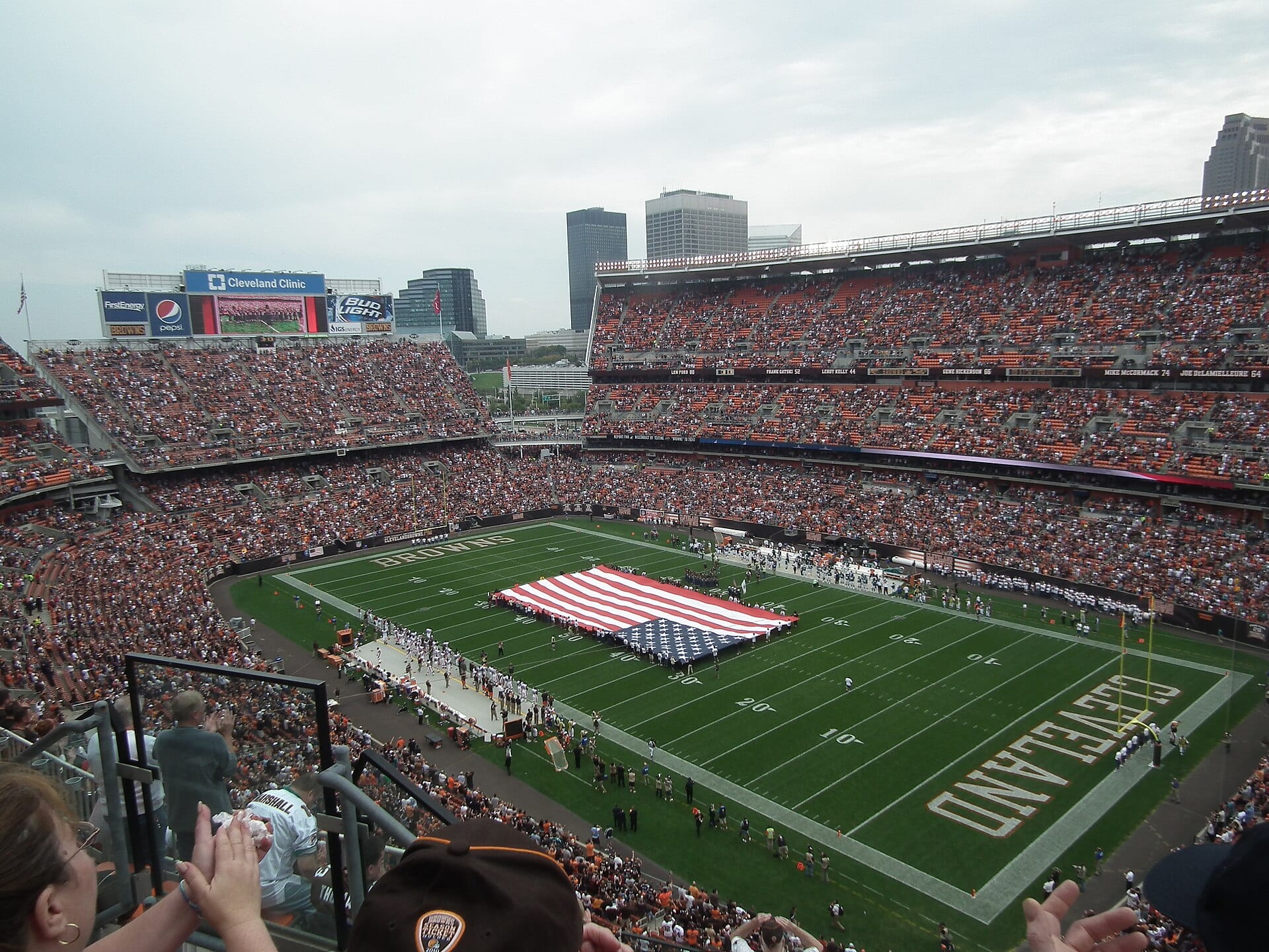 Vista desde la tribuna alta del estadio de los Browns en Cleveland, lleno, con una bandera estadounidense gigante extendida sobre el campo.