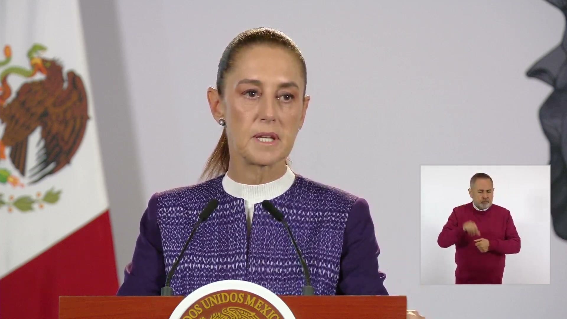 La presidenta Claudia Sheinbaum habla ante dos micrófonos en el podio, con una intérprete de lengua de señas a su derecha.