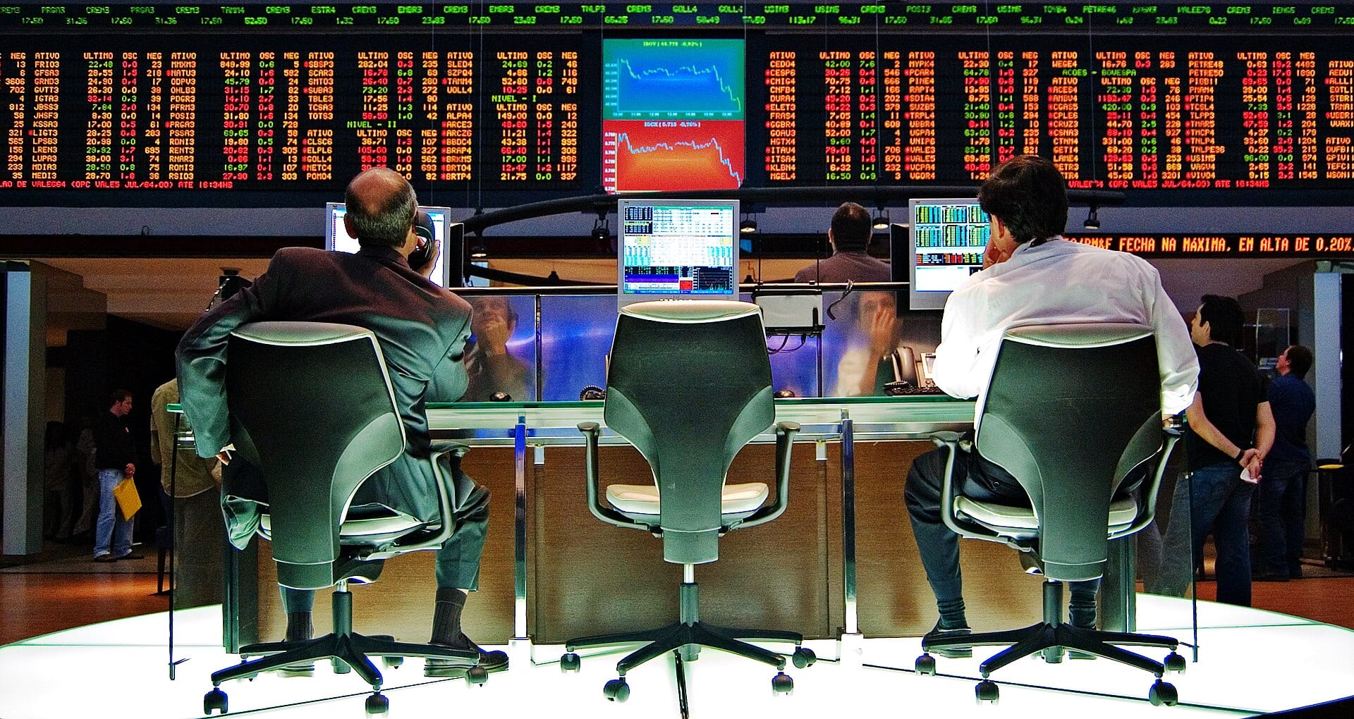 Two traders seated with their backs to the camera in front of a board of green and red quotes and two falling charts, at the São Paulo stock exchange.