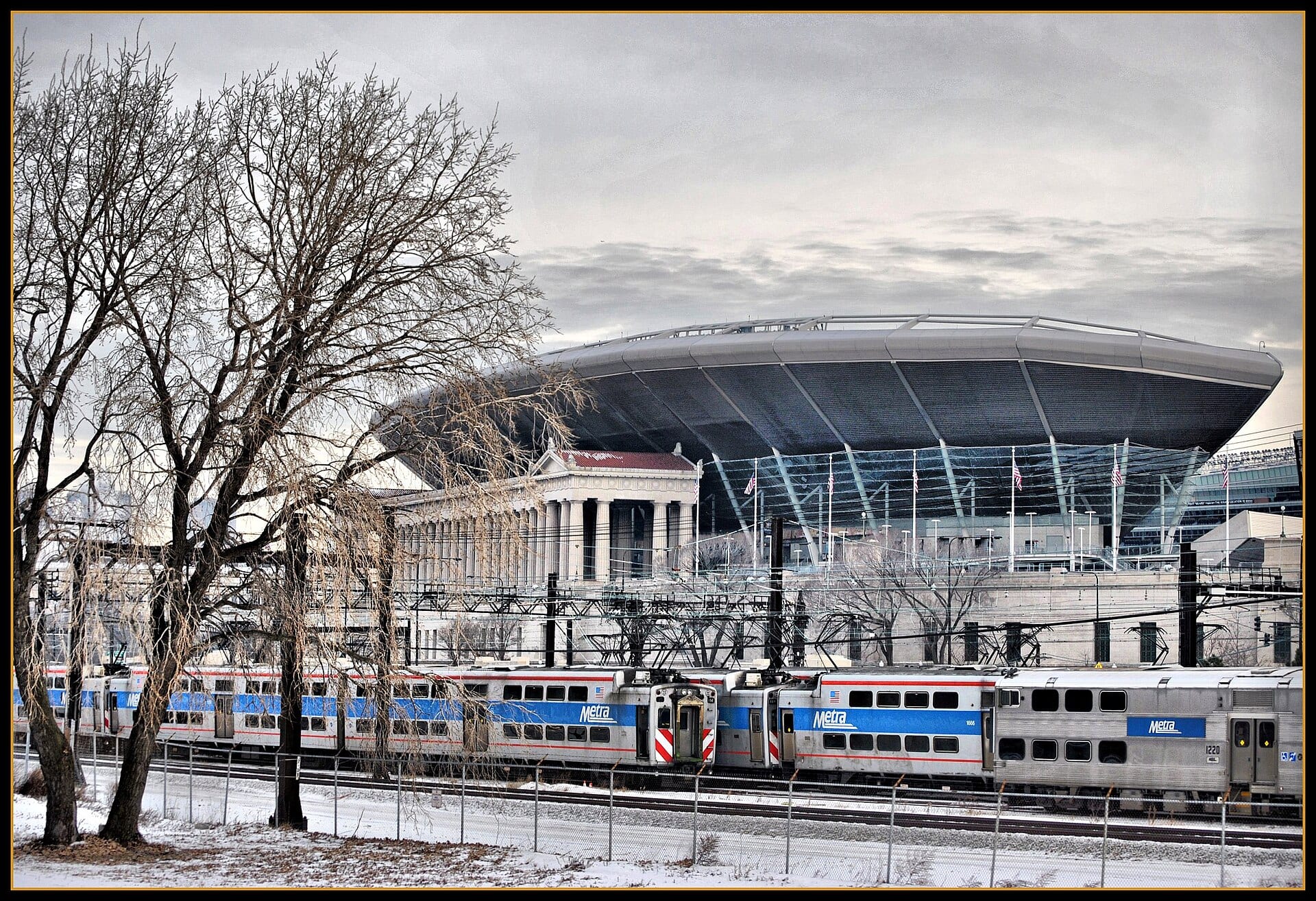 El estadio Soldier Field con su techo moderno sobre la columnata clásica, detrás de un tren Metra y árboles sin hojas.