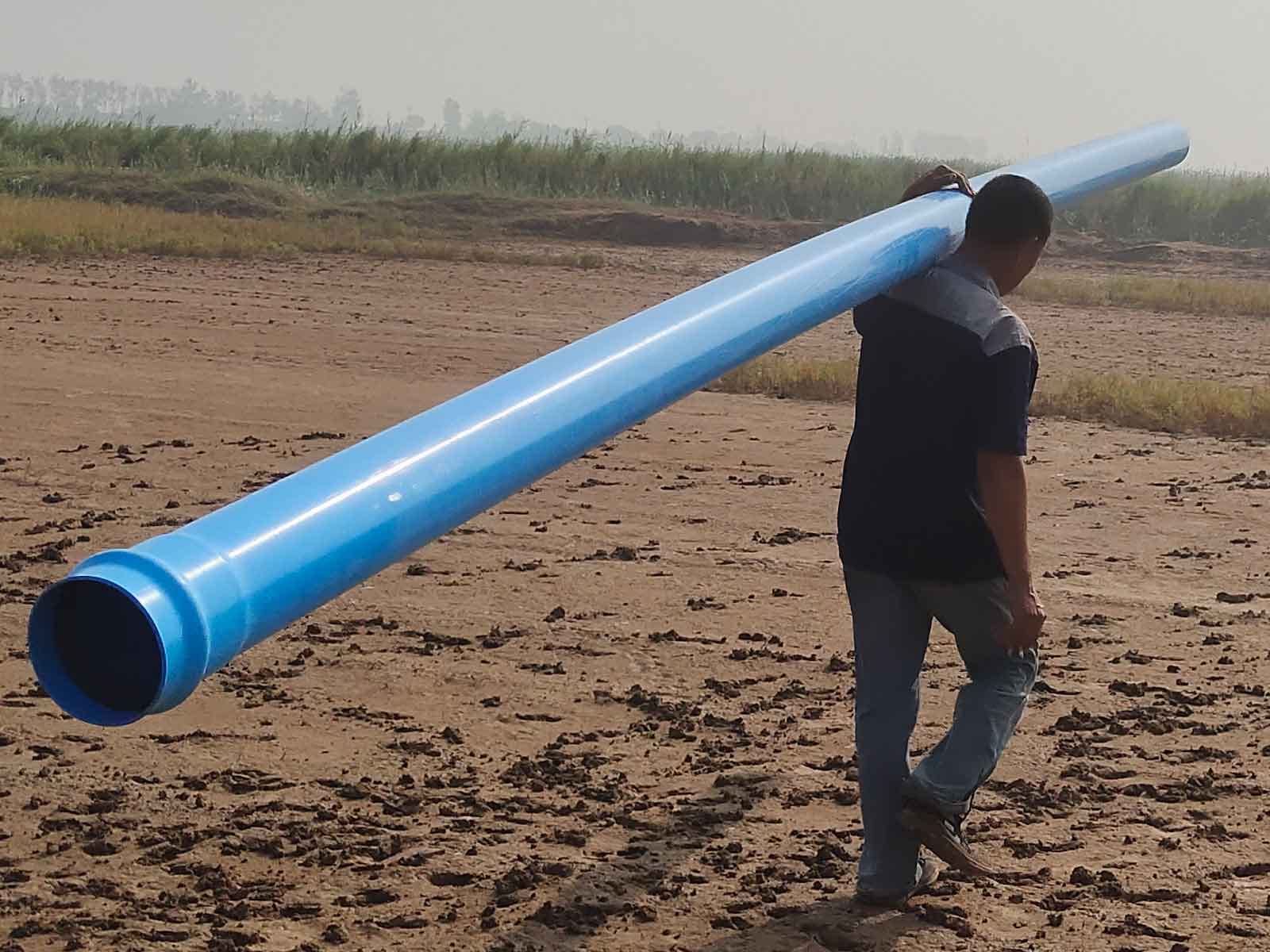 A man, seen from behind, carries a long blue PVC pipe on his shoulder across bare dirt ground.
