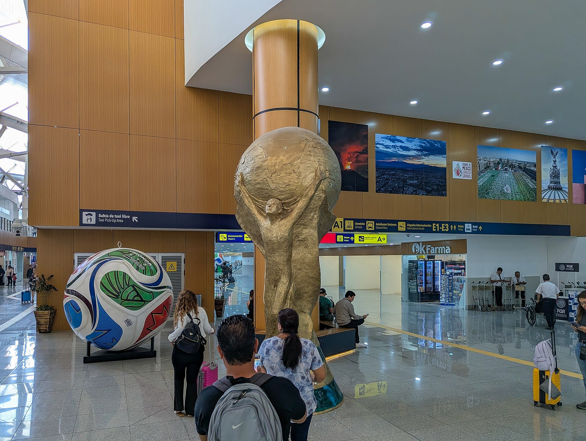 Vestíbulo de una terminal aérea con una escultura dorada de una figura que sostiene un globo, un balón gigante y viajeros con maletas.