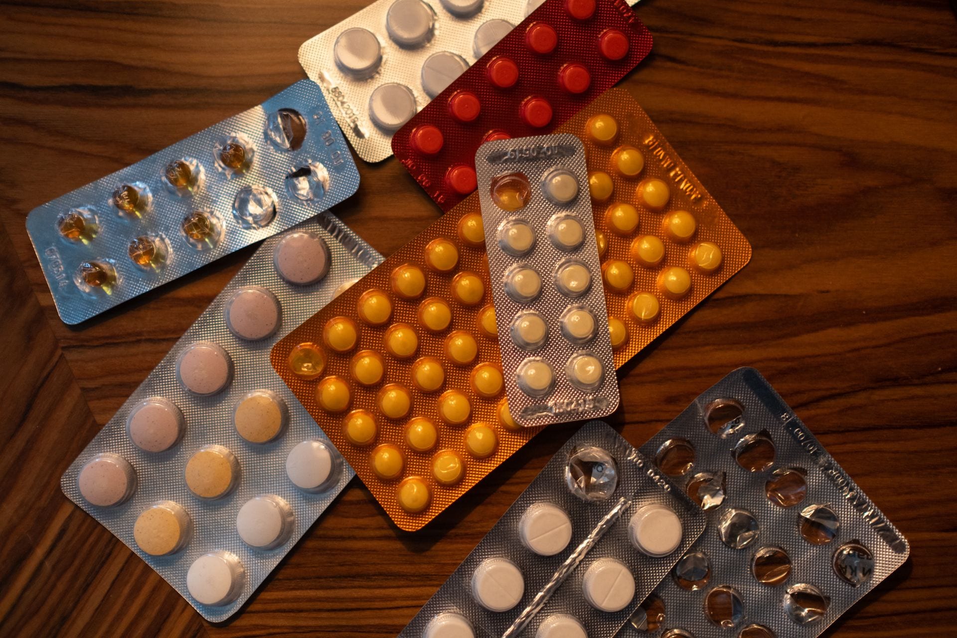 Varias tiras de pastillas en envases de aluminio y plástico, rojas, naranjas, azules y plateadas, sobre una mesa de madera.