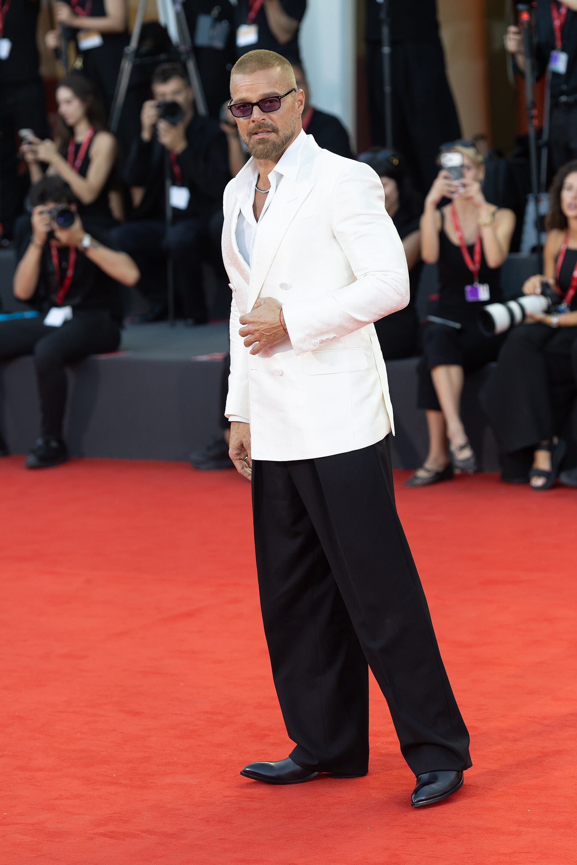 Ricky Martin camina por una alfombra roja con blazer blanco y pantalón negro, rodeado de fotógrafos.