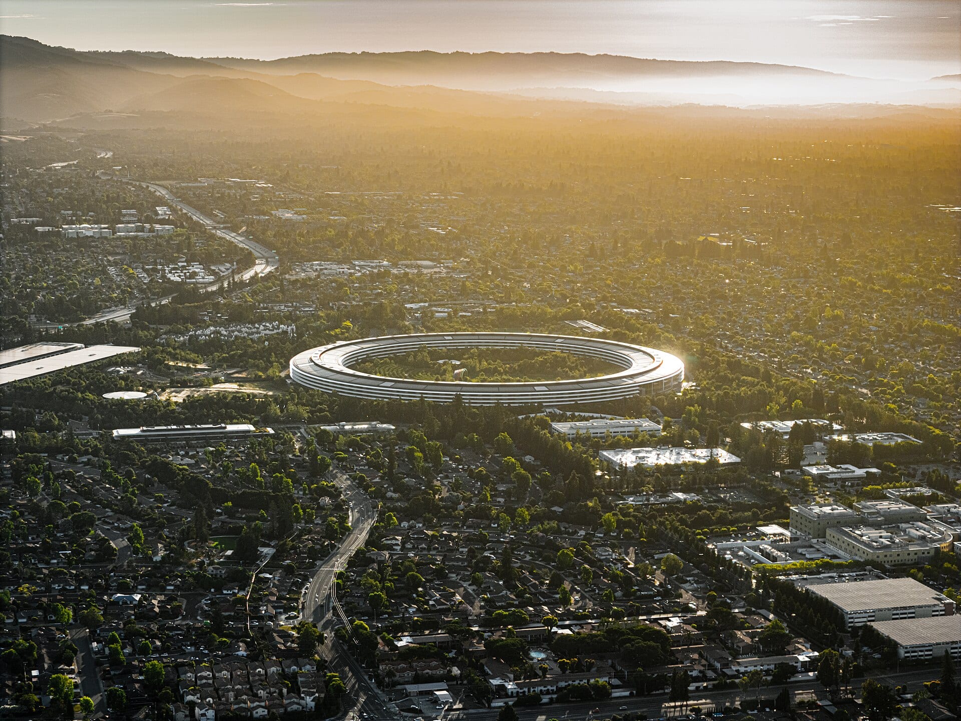 Vista aérea al atardecer del edificio circular de Apple Park, rodeado de árboles y calles residenciales.