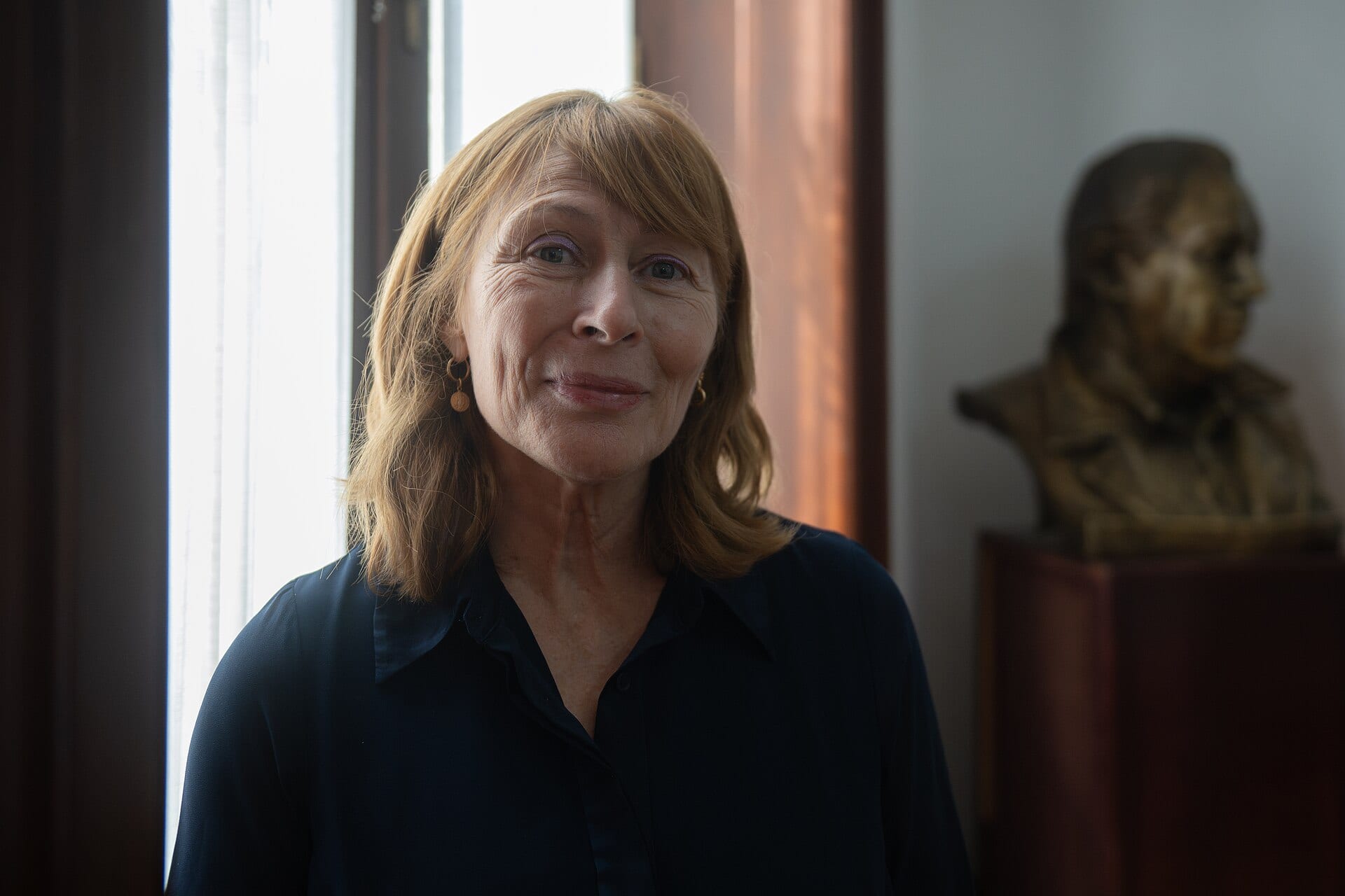 Una mujer de cabello castaño claro y camisa azul oscuro sonríe junto a una ventana, con un busto de bronce al fondo.