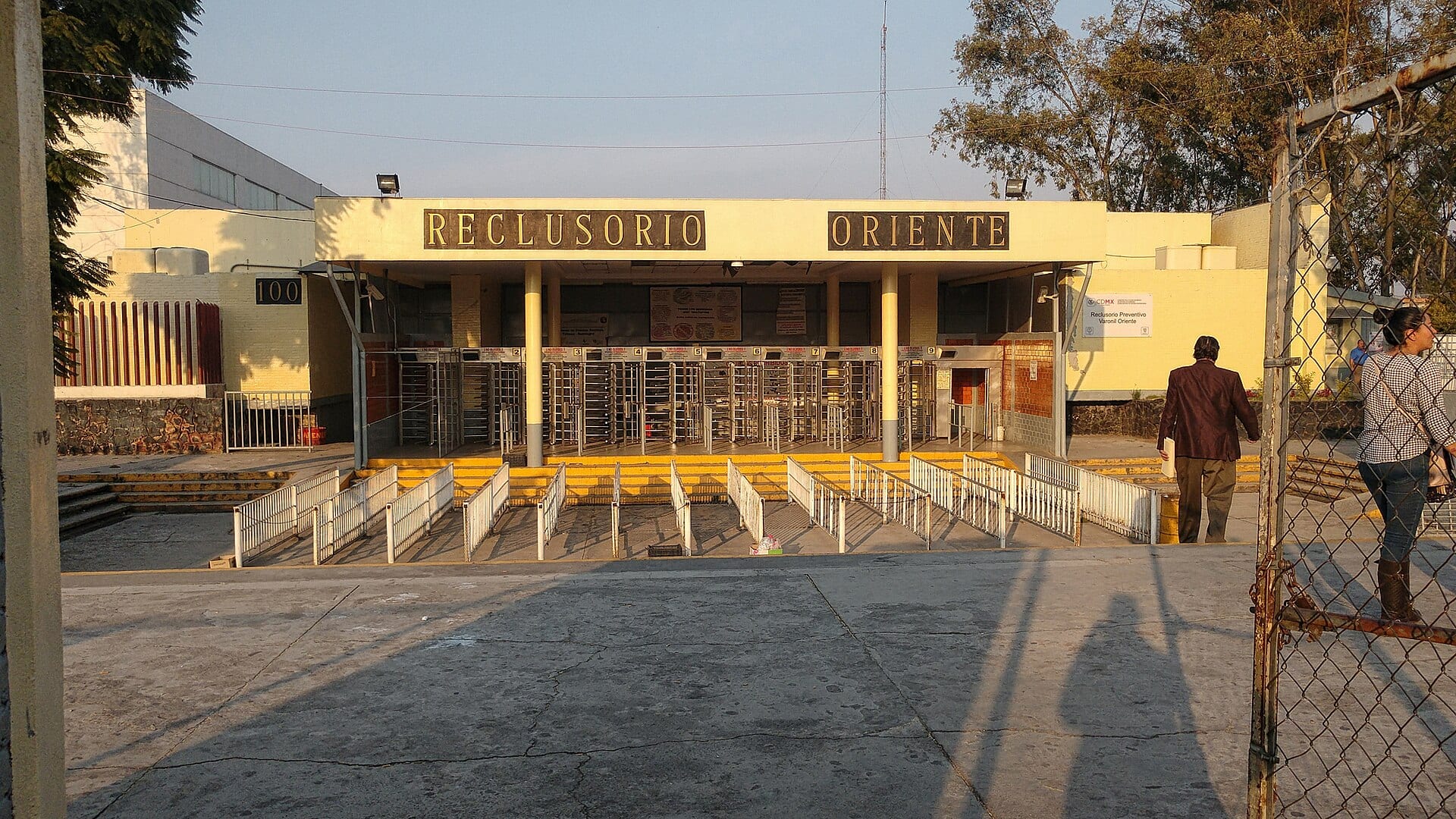 Fachada amarilla del acceso al Reclusorio Oriente, con torniquetes, barandales blancos y dos personas a la derecha del encuadre.