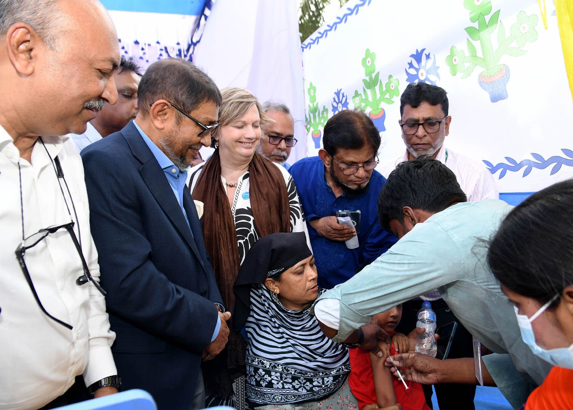 A health worker gives an injection in the arm of a child sitting beside his mother, surrounded by officials, in a vaccination tent.