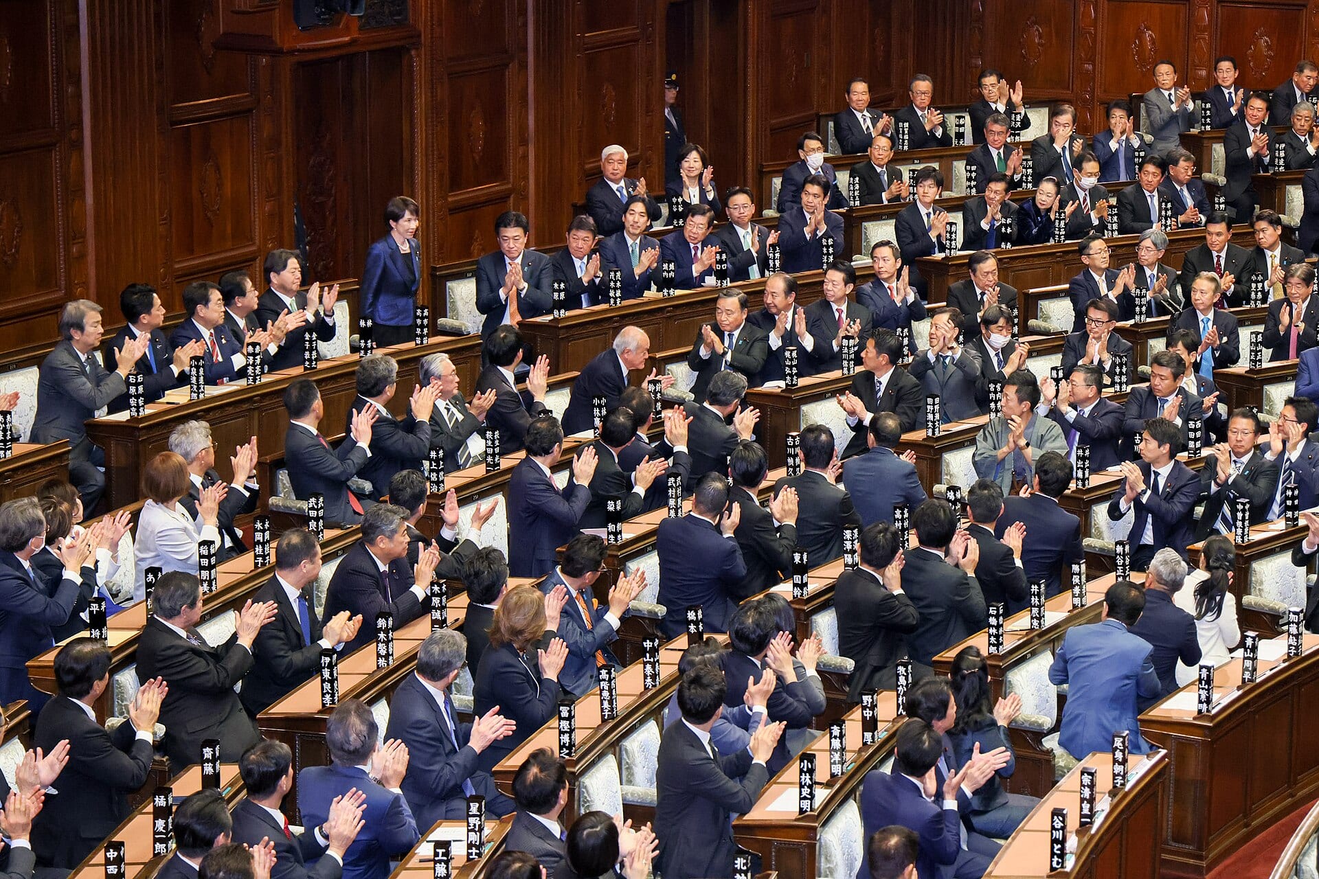 A woman in a blue suit stands in the chamber of a legislature while dozens of lawmakers in dark suits applaud from their seats.