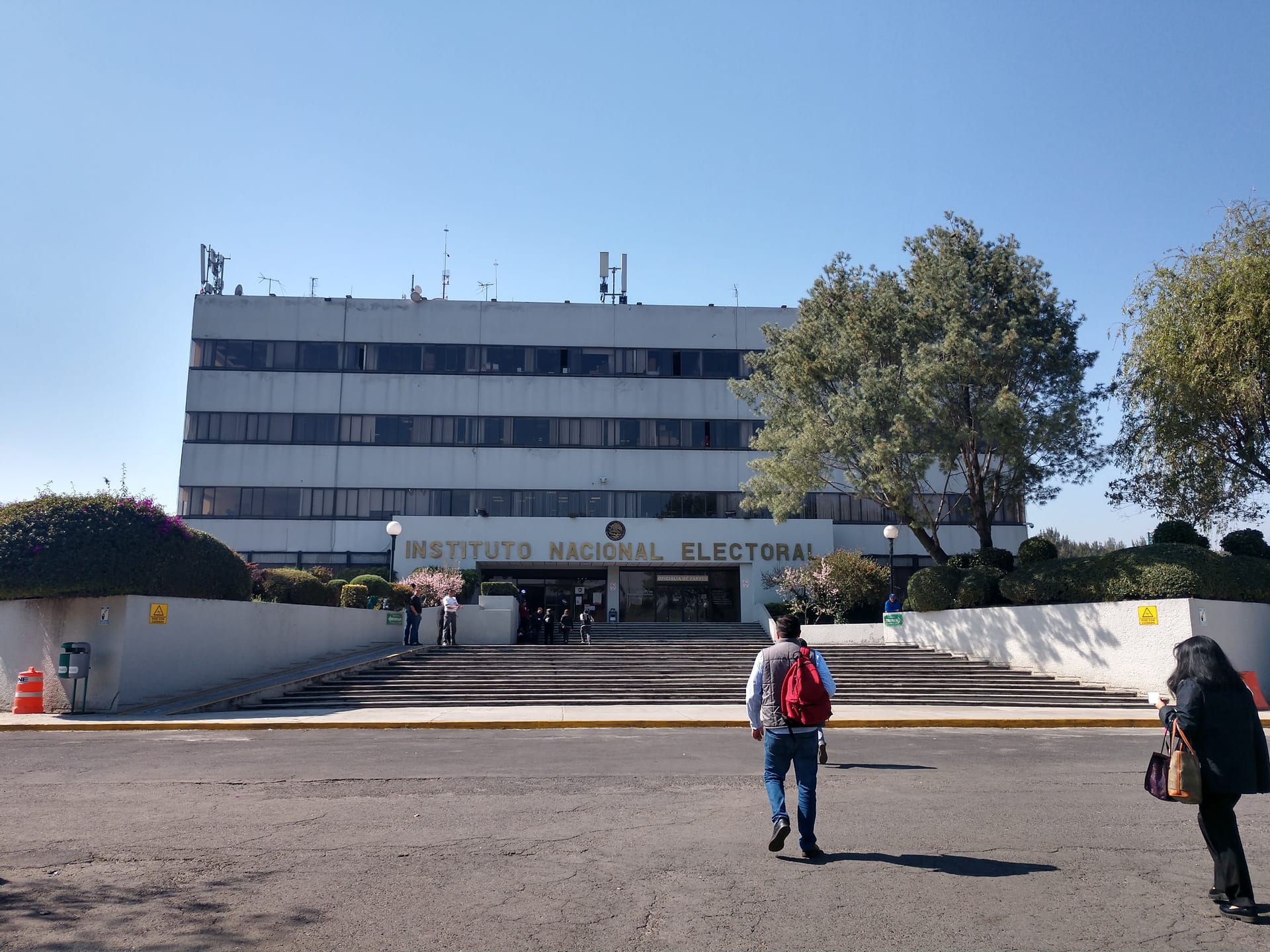 Edificio de varios pisos con el letrero "Instituto Nacional Electoral" en la fachada, entre árboles y personas que caminan hacia la entrada.