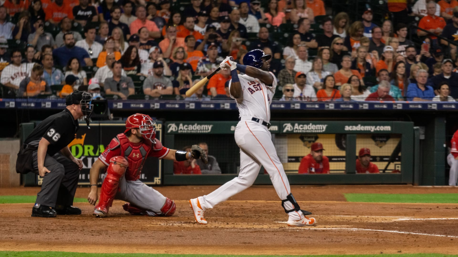 Yordan Álvarez, con casco azul marino y uniforme blanco de los Astros, termina un swing frente al cátcher y el árbitro de home, con las gradas llenas al fondo.