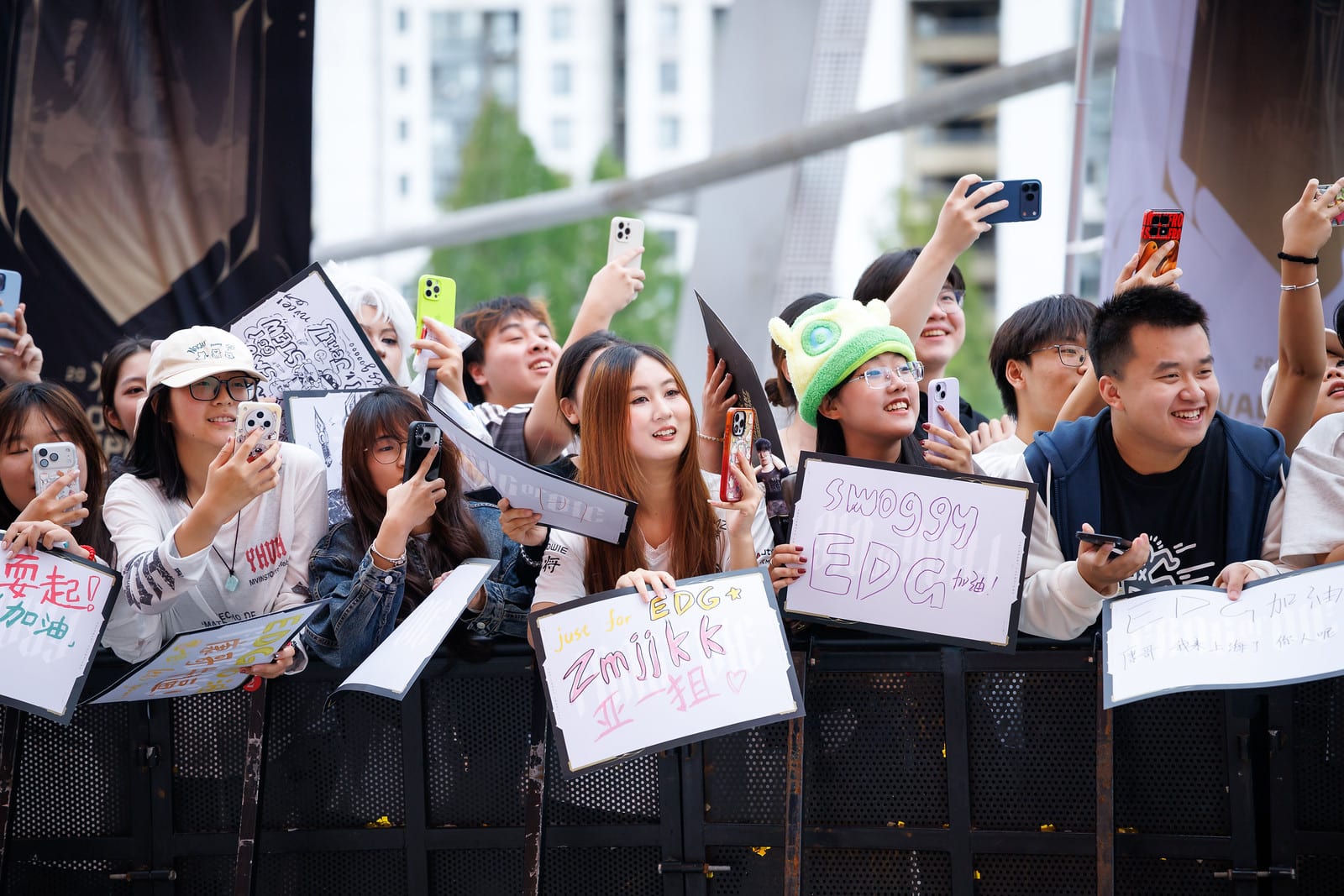 Fans crowd behind a barrier with handmade signs reading EDG, under the metal structure of the venue.