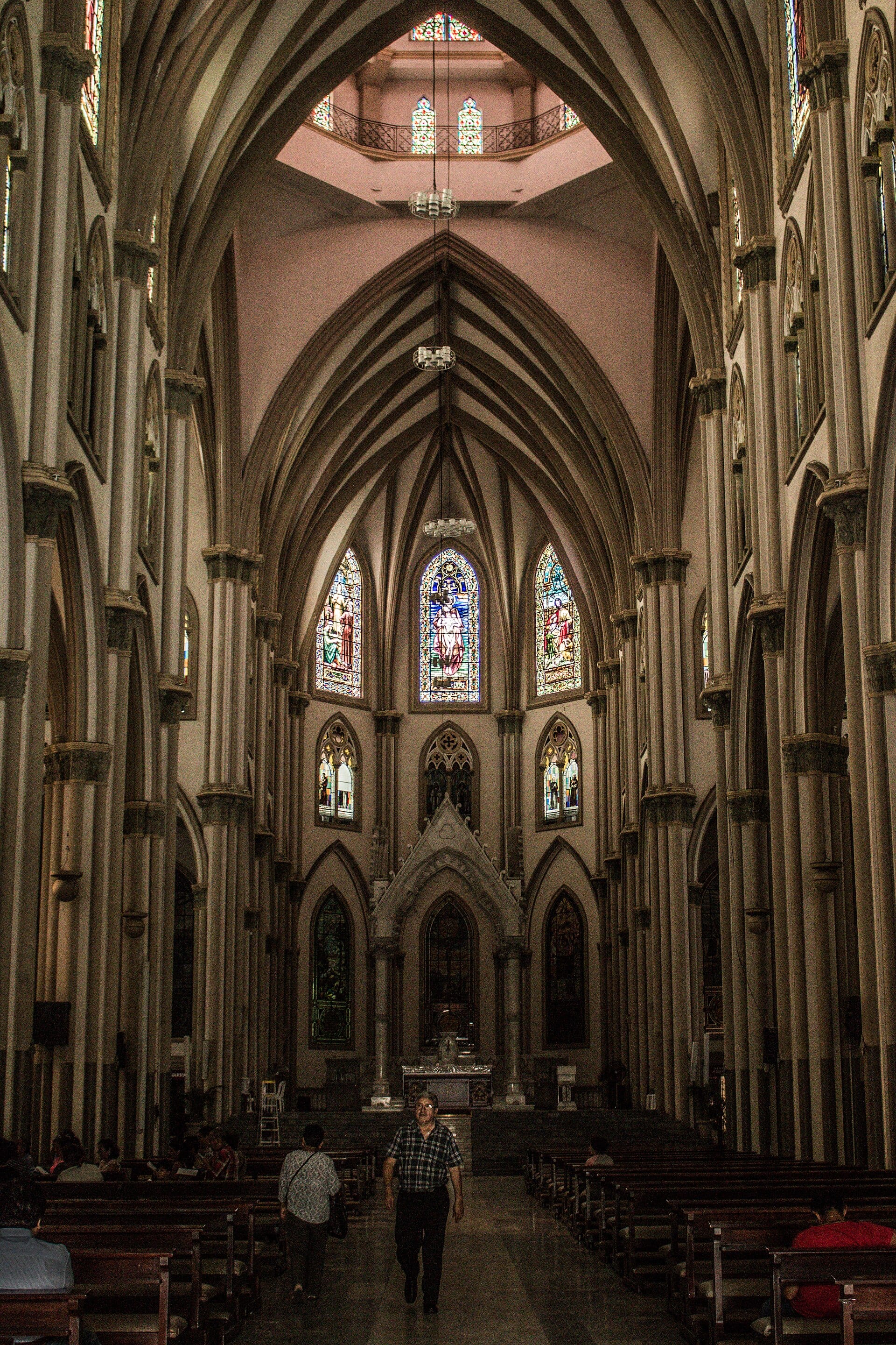 Nave central de una catedral gótica con vitrales al fondo y bancas de madera; algunas personas caminan por el pasillo.