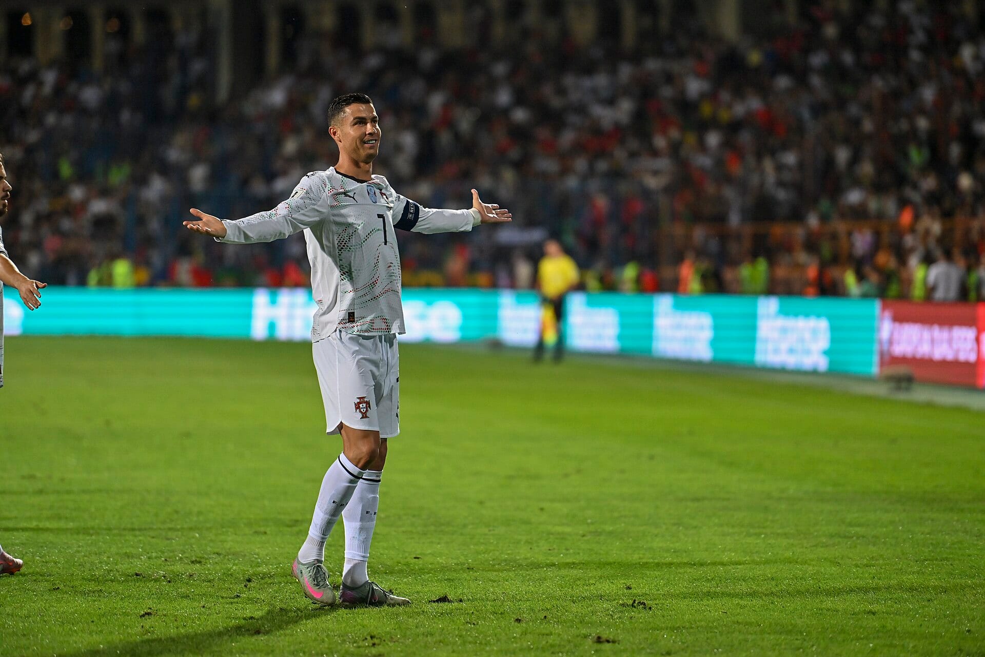 Cristiano Ronaldo, in a white Portugal shirt and captain's armband, spreads his arms on the pitch in front of a full stand.
