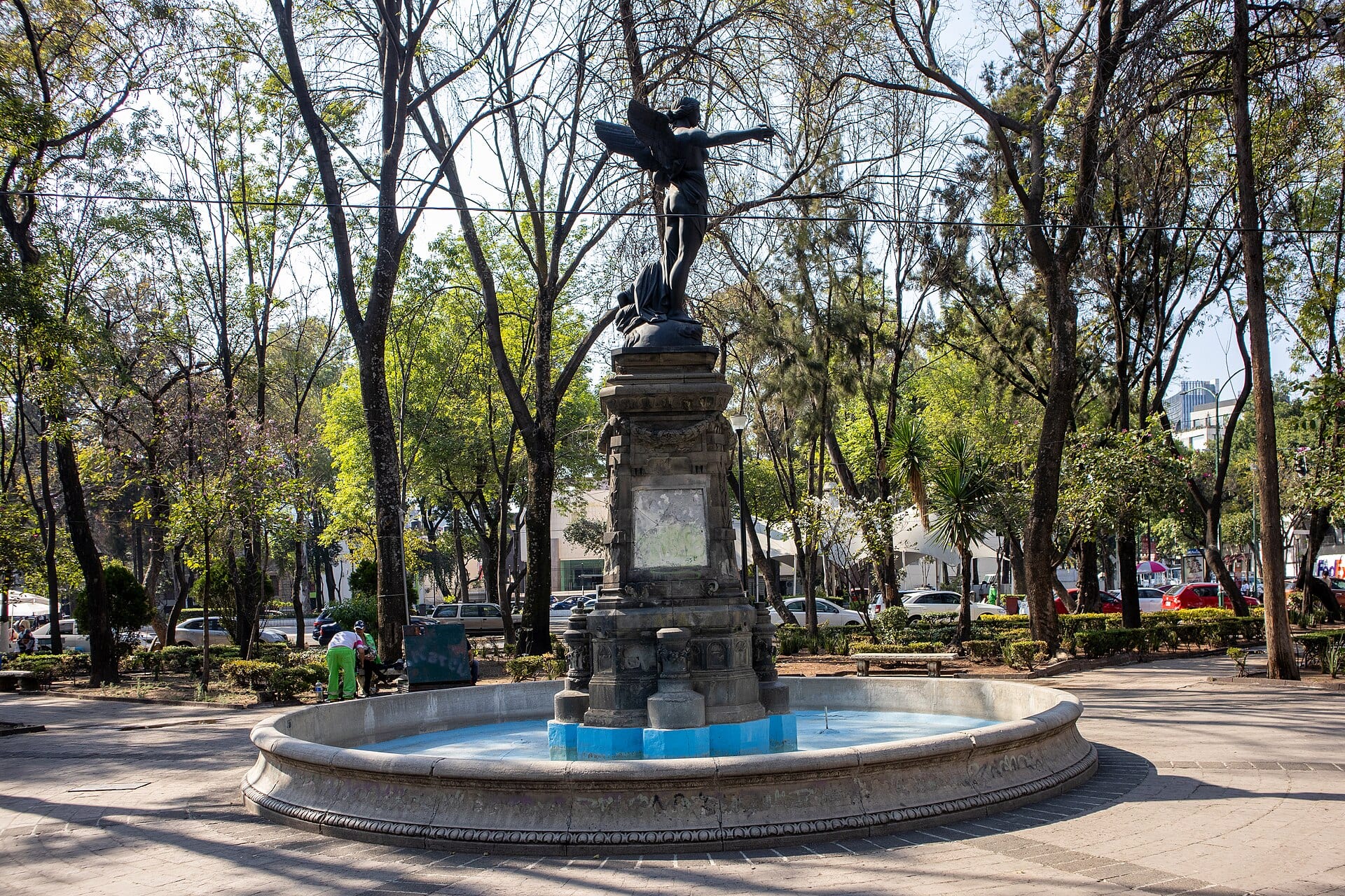 Una fuente circular con una escultura alada sobre un pedestal de piedra, rodeada de árboles en un parque de la Ciudadela.