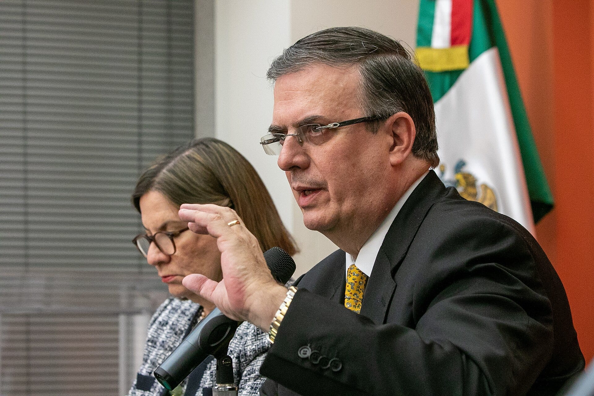 Marcelo Ebrard, con lentes y traje oscuro, habla ante un micrófono junto a una bandera de México.