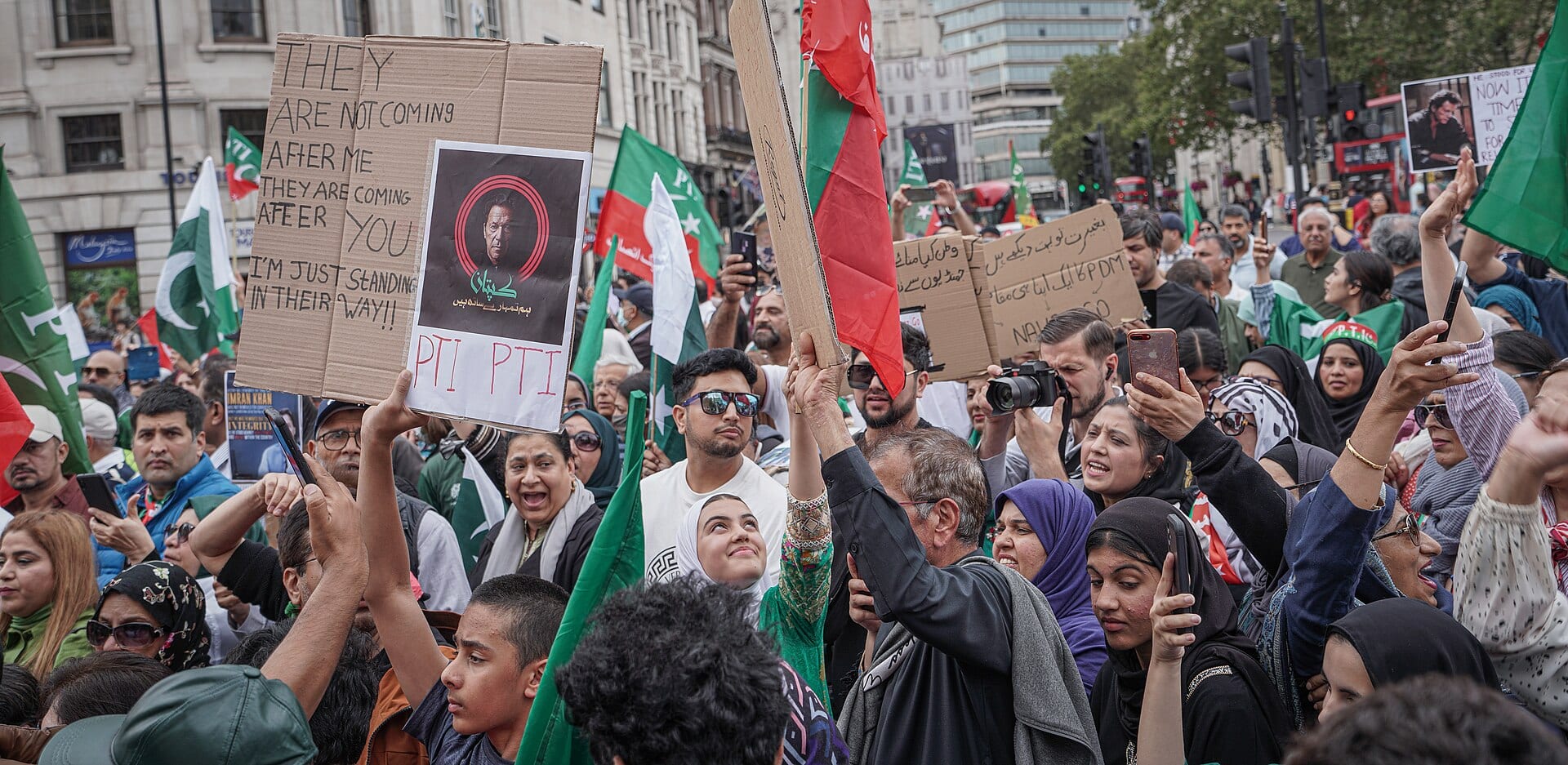 Protesters with green flags and signs, one bearing a portrait of Imran Khan, crowd together at a 2023 demonstration in London.