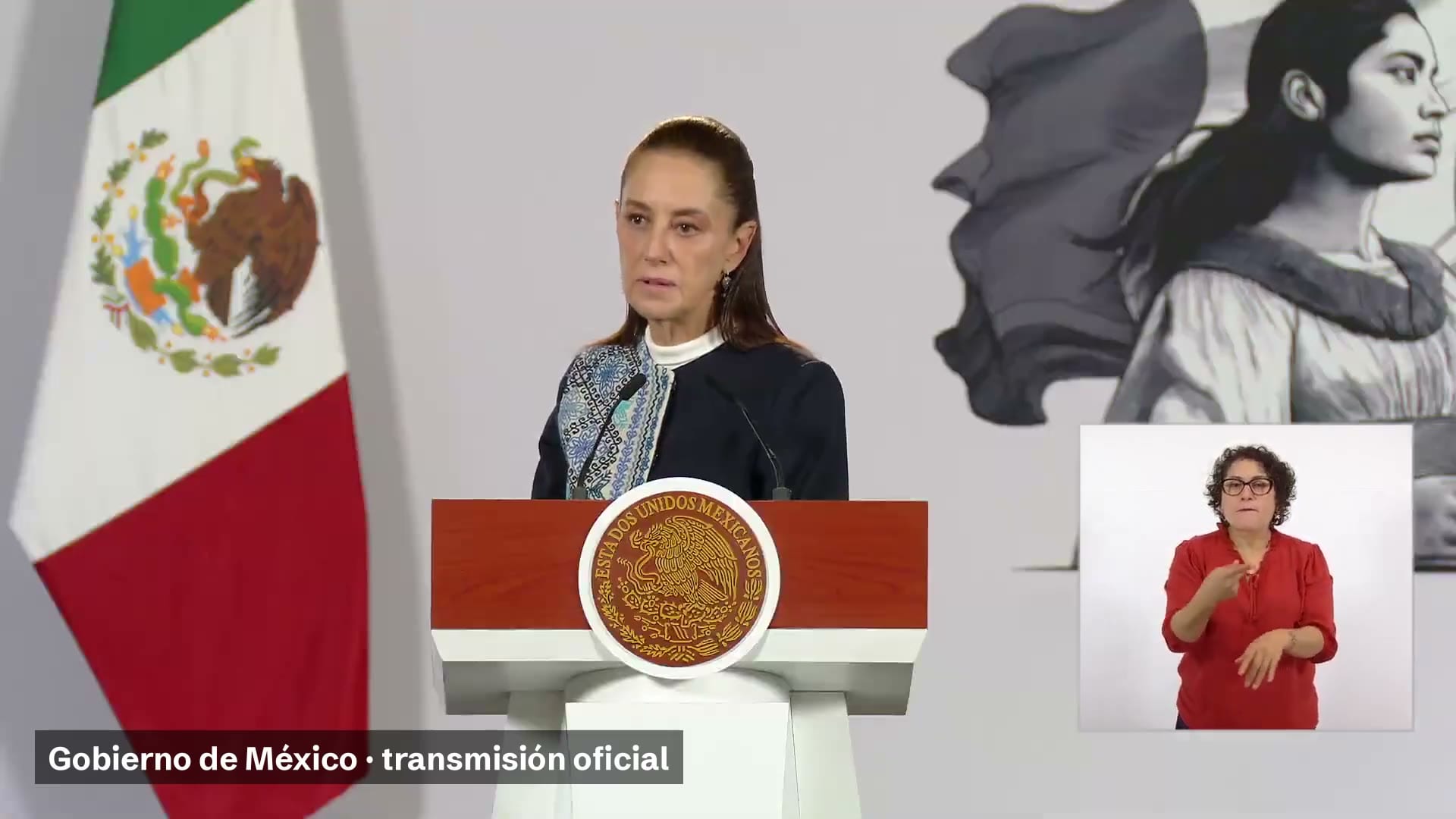 La presidenta Claudia Sheinbaum habla frente a un podio con el escudo nacional, junto a una bandera de México y una intérprete de lengua de señas.
