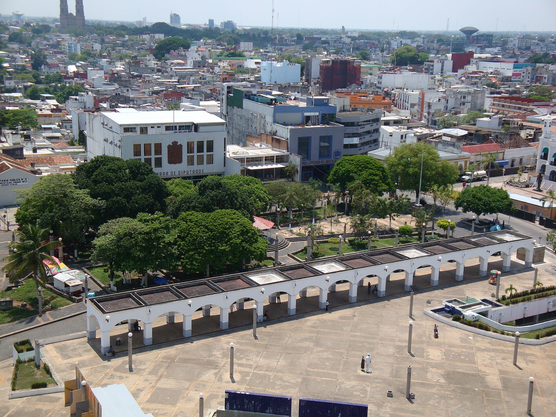 Vista aérea de la Plaza de Armas de Villahermosa, con un portal de arcos blancos, árboles y, al fondo, los edificios del Tribunal Superior de Justicia y del Congreso estatal.