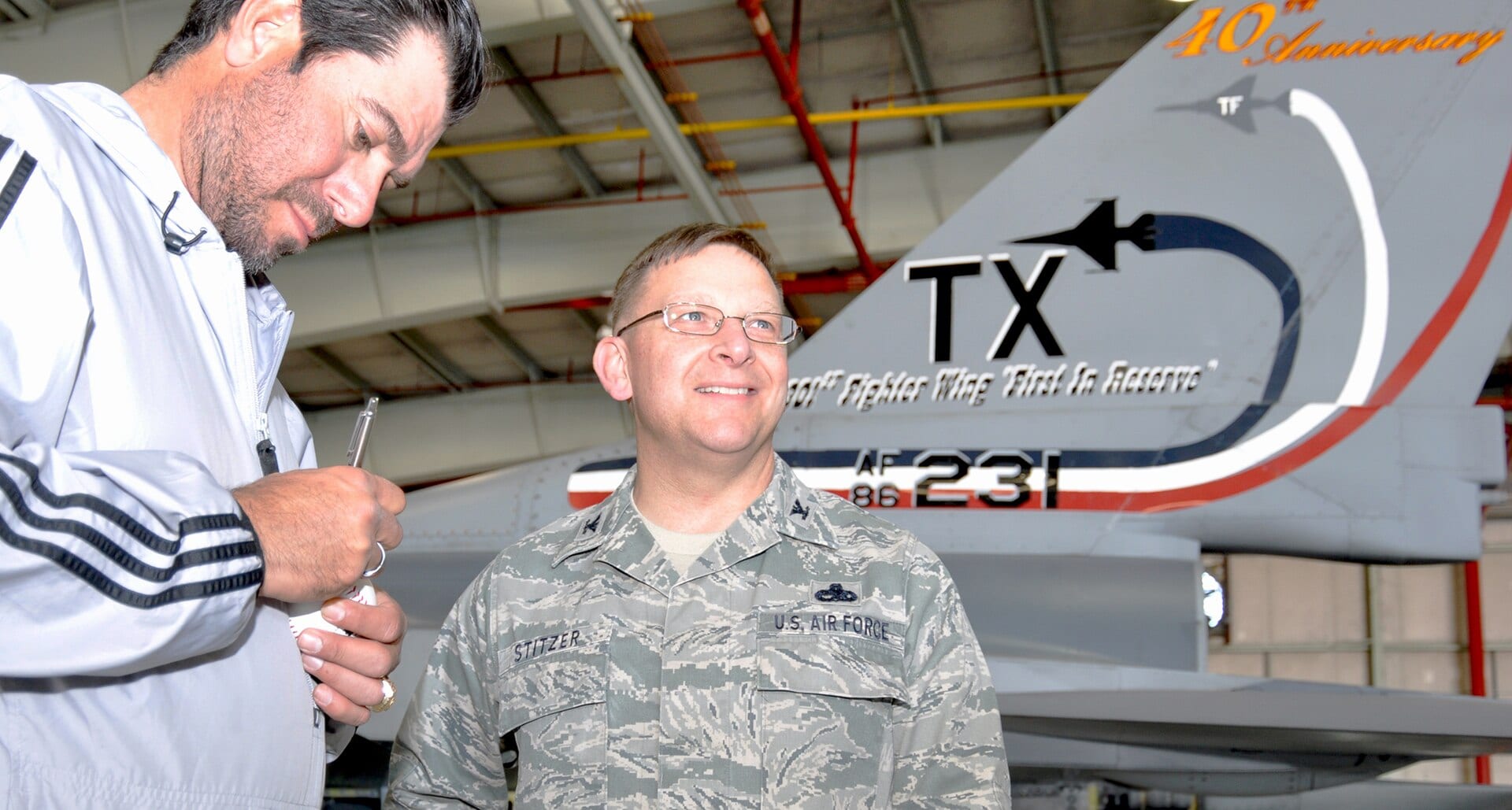 Benjamín Gil, de chamarra gris, firma una pelota de beisbol junto a un coronel de la Fuerza Aérea frente a la cola de un caza, en un hangar.