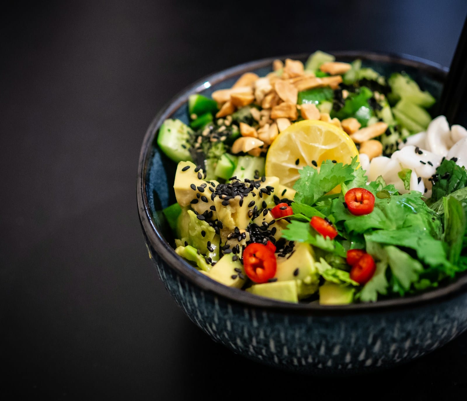Un tazón azul oscuro con aguacate en cubos, pepino, cacahuates, medio limón, cilantro y chile rojo sobre fondo oscuro.