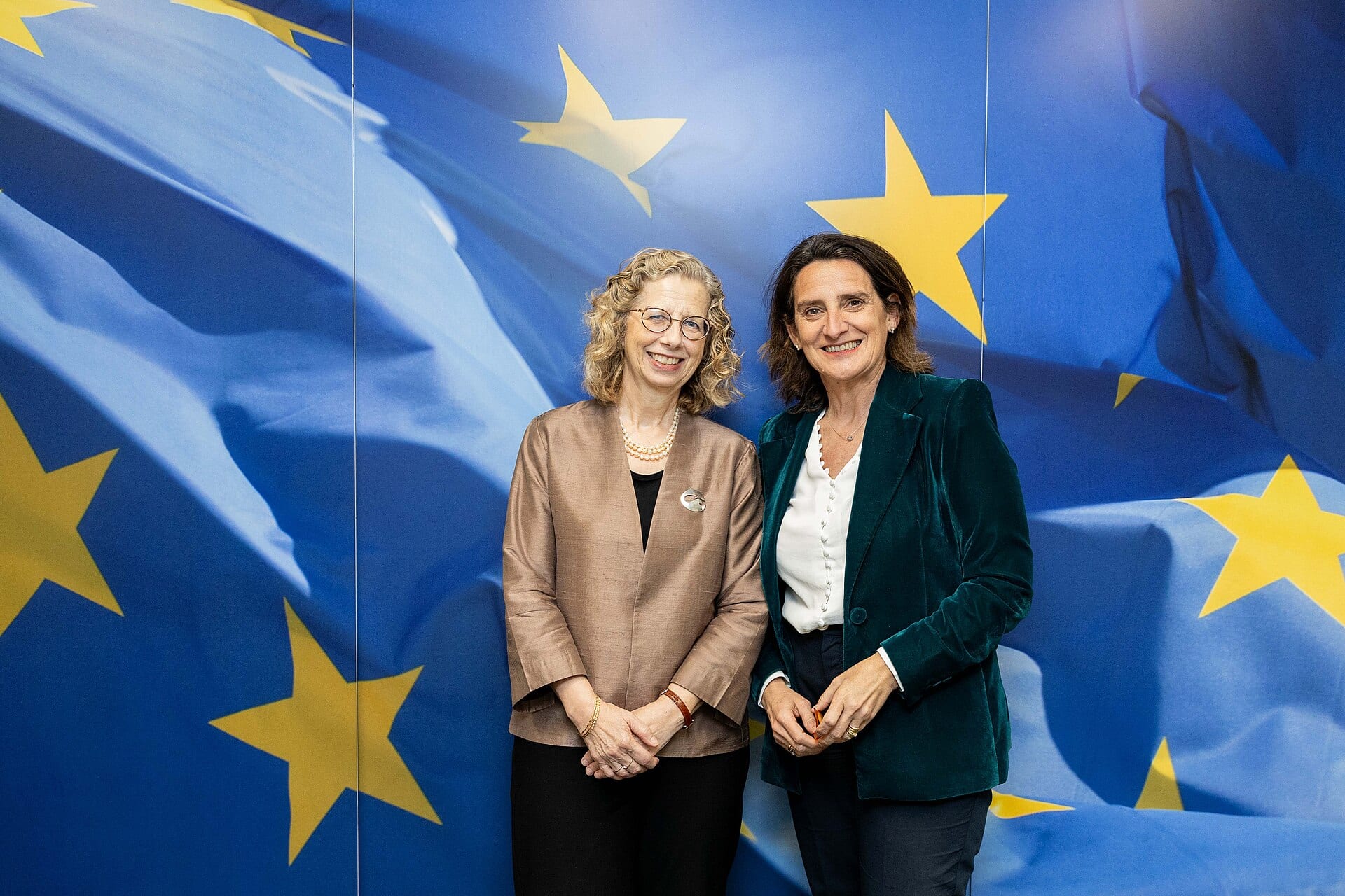 Inger Andersen, a la izquierda, sonríe junto a otra mujer frente a un fondo azul con las estrellas amarillas de la bandera europea.