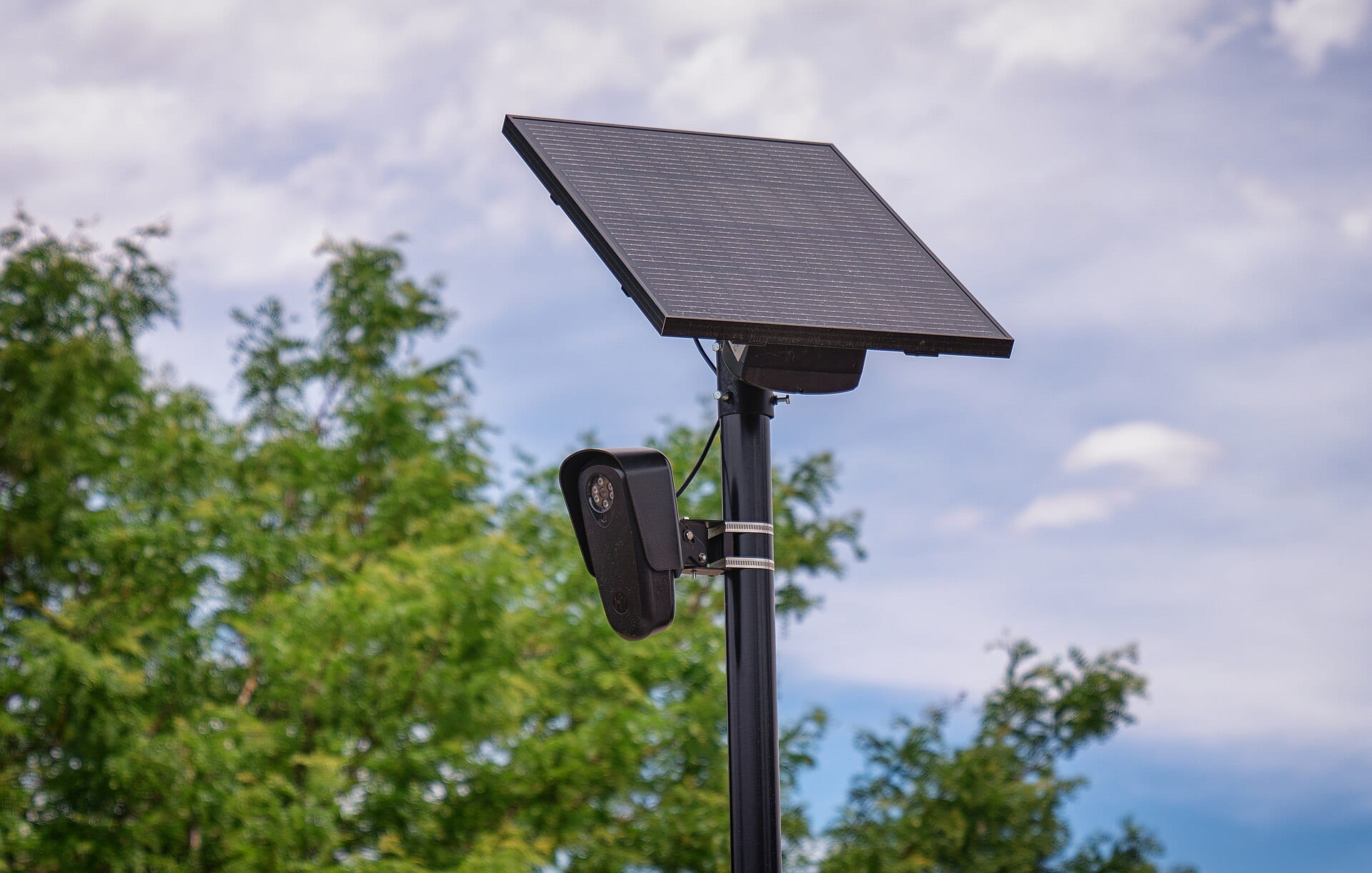 Una cámara negra de lectura de placas montada en un poste, con un panel solar inclinado encima y árboles al fondo.