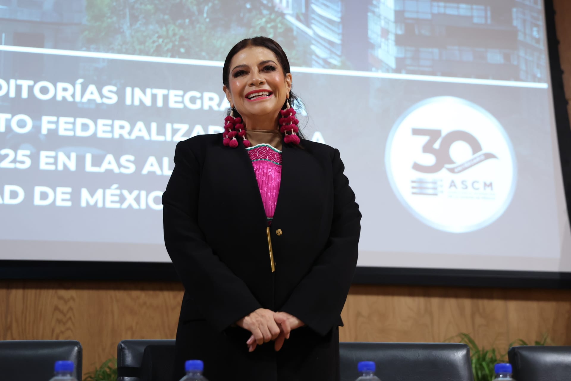 Una mujer de saco negro sonríe de pie frente a una pantalla con el logotipo de la Auditoría Superior de la Ciudad de México.