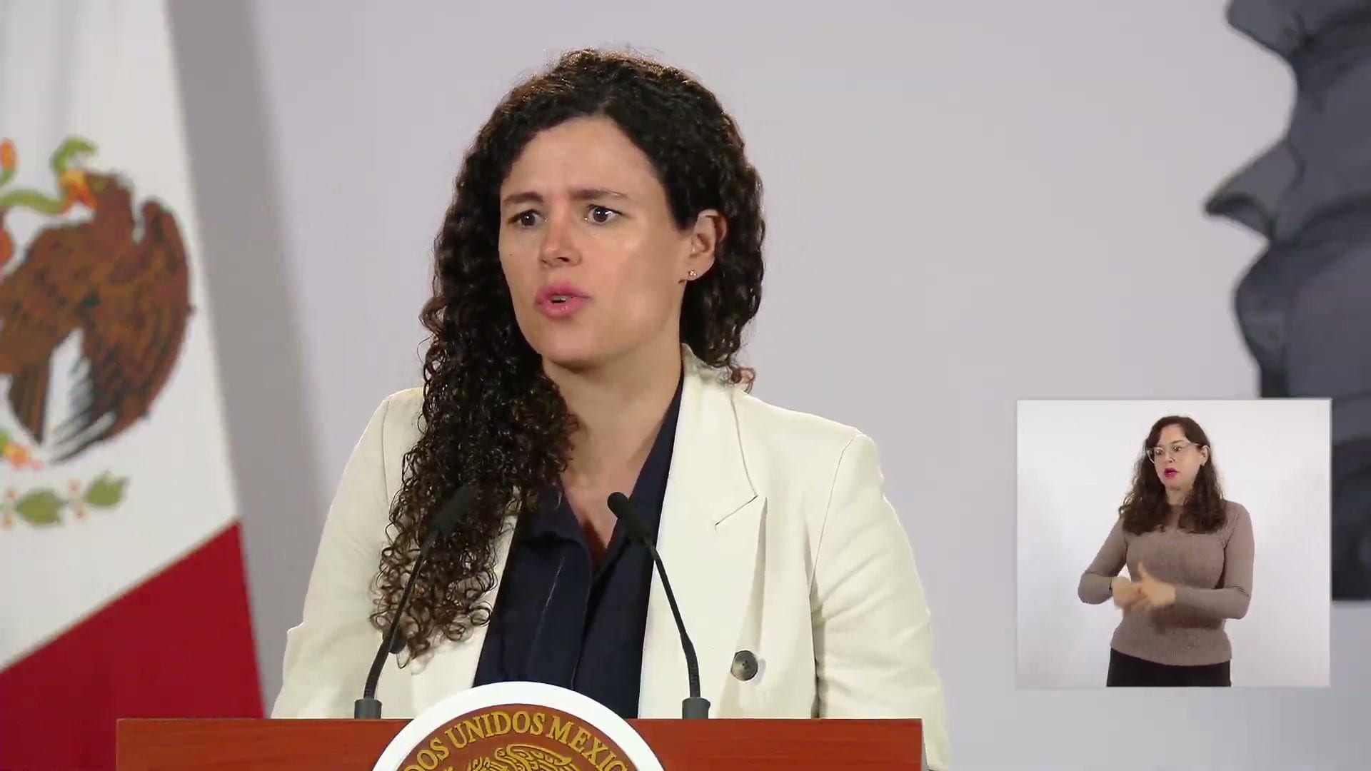 Una mujer de cabello rizado y saco blanco habla detrás de un podio con el escudo nacional, junto a una bandera de México.