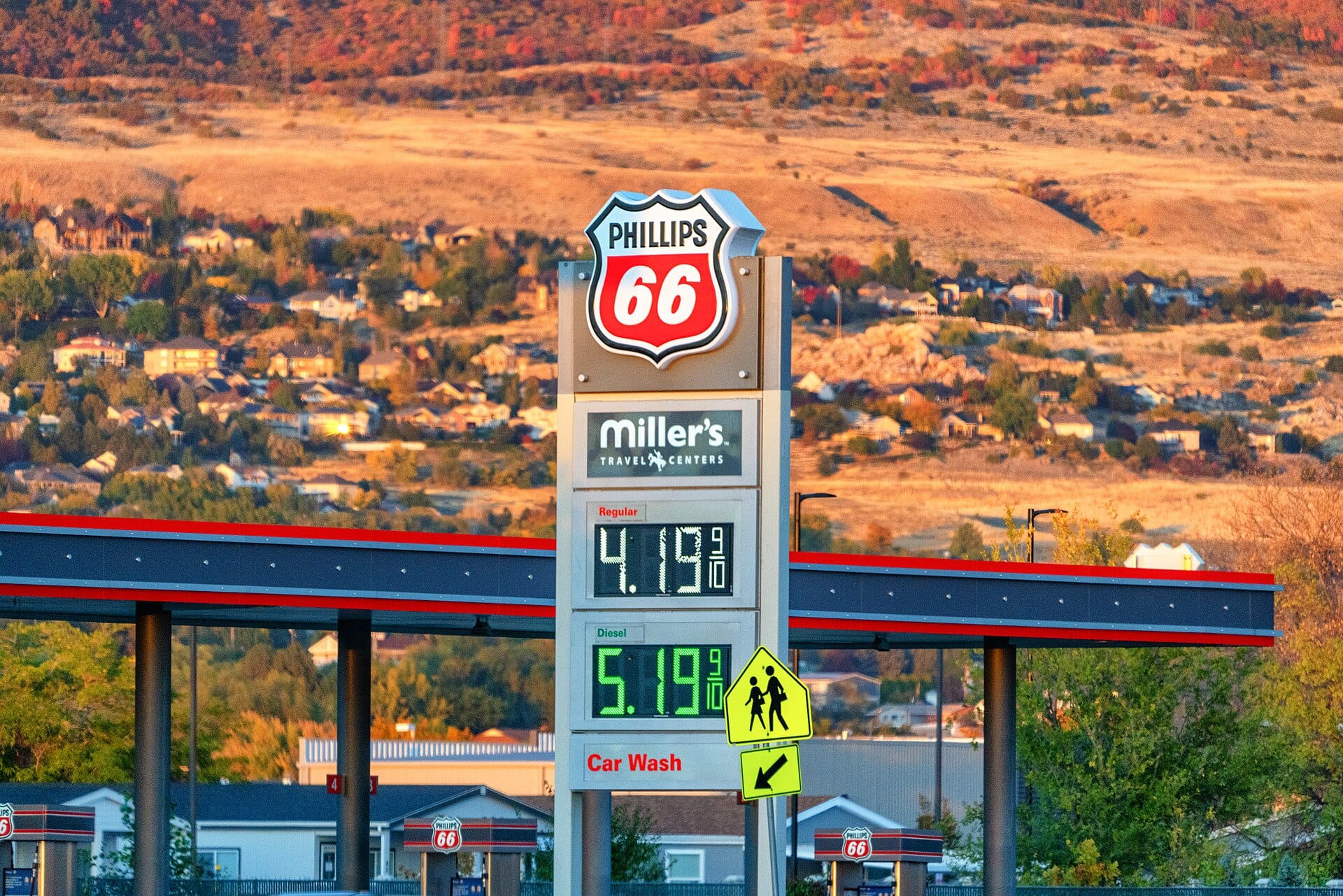 A Phillips 66 gas station sign shows regular and diesel prices on lit displays, in front of a hillside with houses.