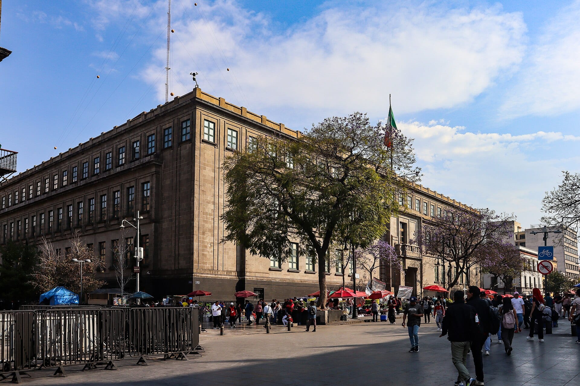 Esquina del edificio de piedra de la Suprema Corte, con un árbol al frente, vallas metálicas y peatones cruzando la plaza.
