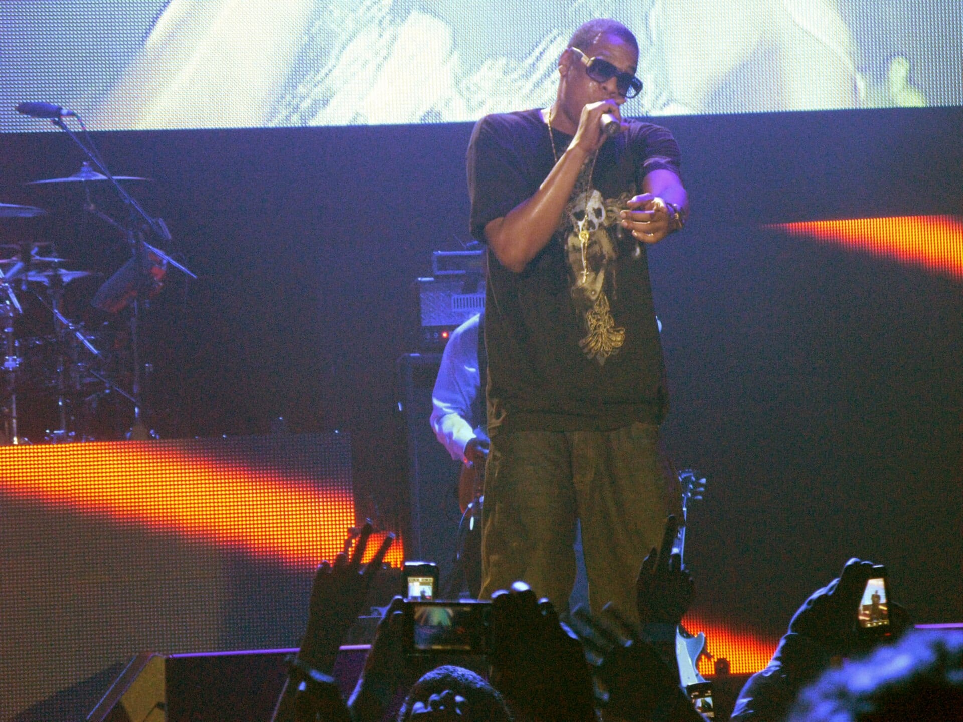 Jay-Z, in dark sunglasses and holding a microphone, points at the crowd from a stage lit by video screens.
