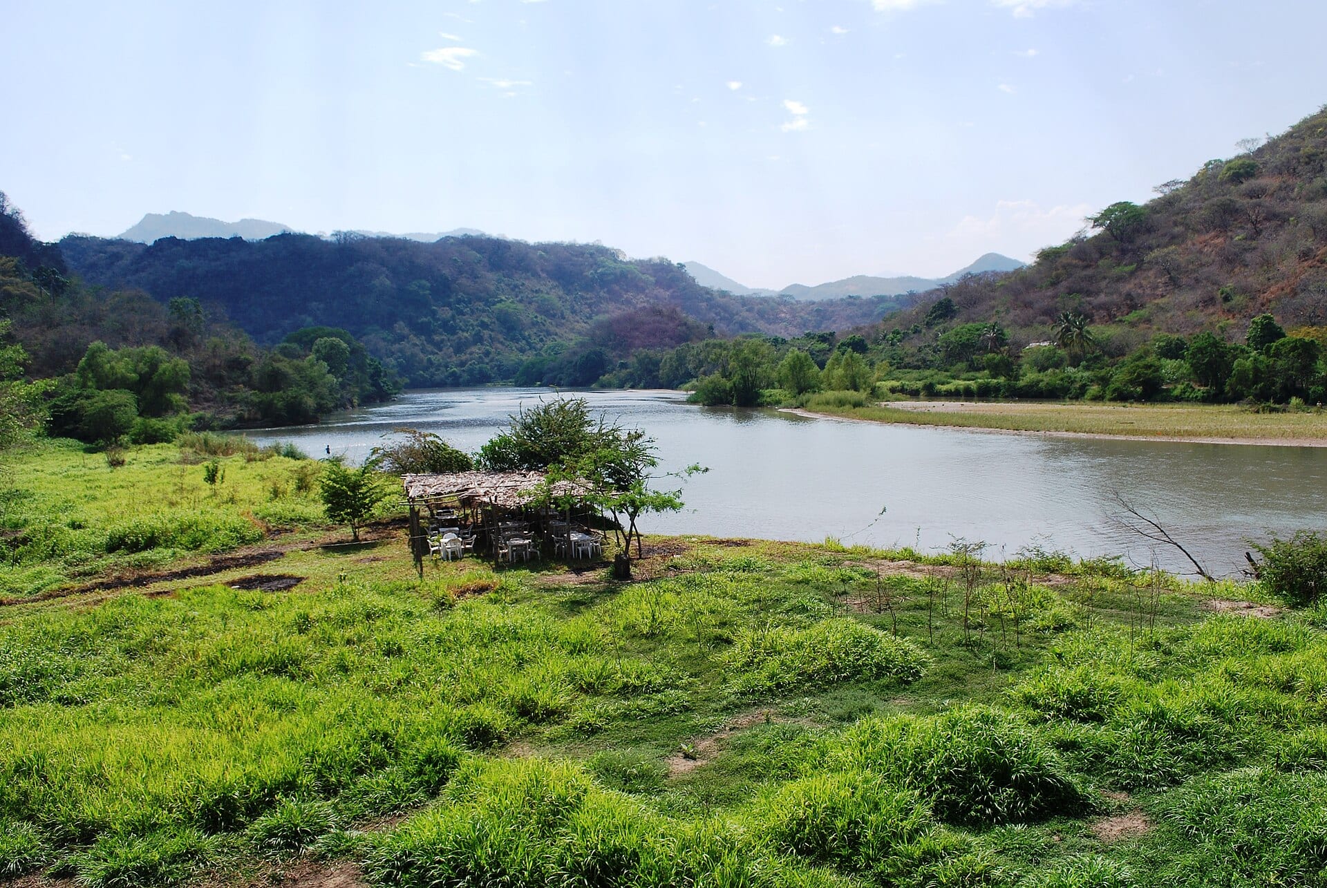Un río de aguas tranquilas entre cerros cubiertos de vegetación, con una enramada de palma y mesas blancas en la orilla cubierta de pasto.
