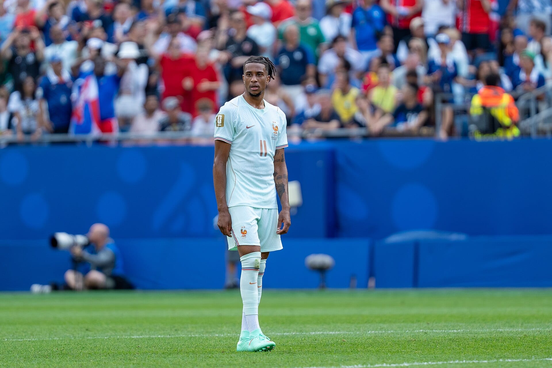 Michael Olise, con la camiseta blanca de Francia y el número 11, de pie sobre el césped con la grada llena detrás.