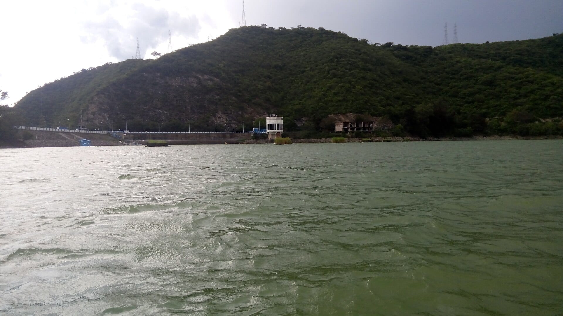 Una cortina de presa con una torre de toma de agua al pie de un cerro boscoso, y agua verde con oleaje en primer plano.