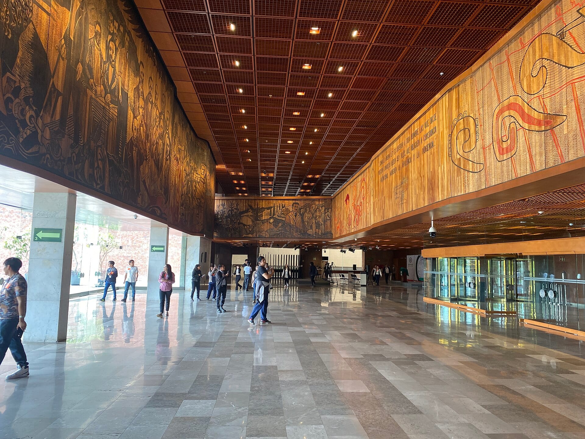 Vestíbulo amplio del Palacio Legislativo de San Lázaro, con murales de madera tallada en los muros y varias personas caminando sobre piso de mármol.