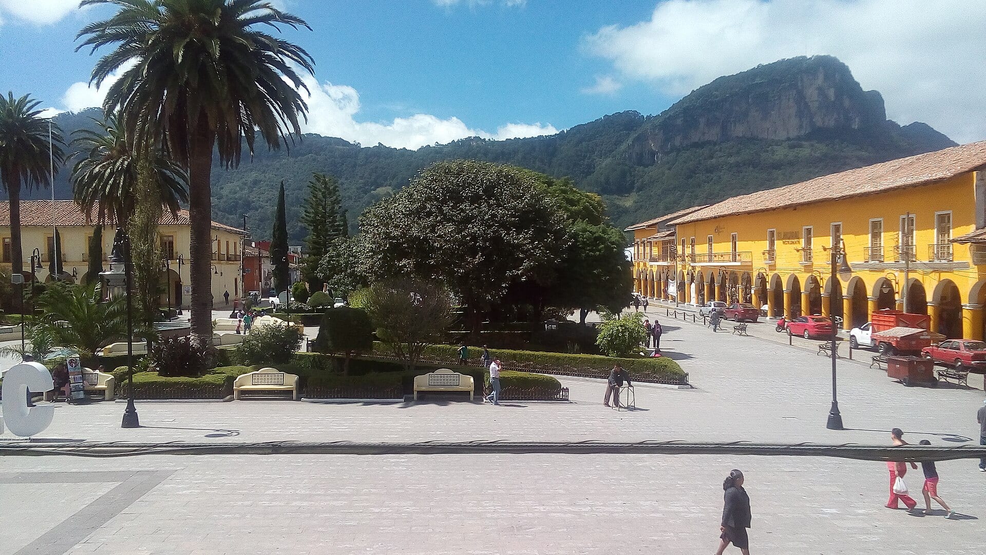Plaza con jardín y palmeras frente a portales amarillos, con un cerro de roca y bosque al fondo.