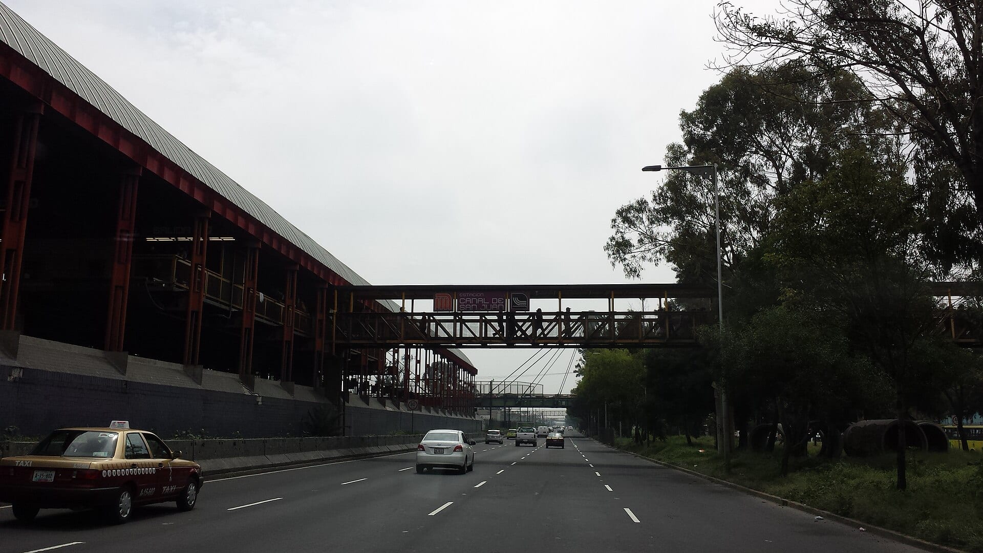 Una avenida de varios carriles con un taxi y autos, junto a la estación Canal de San Juan del Metro, con un puente peatonal que cruza la vialidad entre árboles.