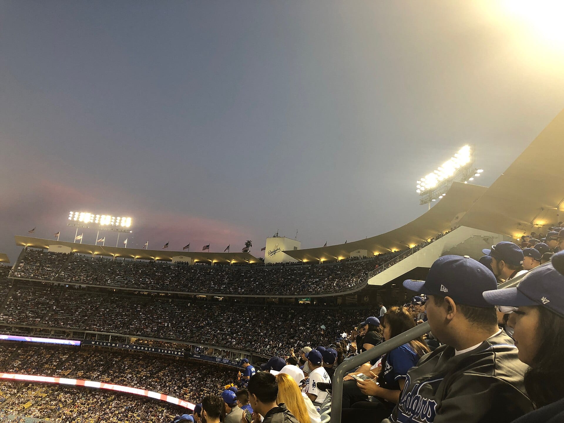 Aficionados con gorras azules en las gradas del Dodger Stadium al anochecer, con las torres de luces encendidas y el letrero de los Dodgers al fondo.