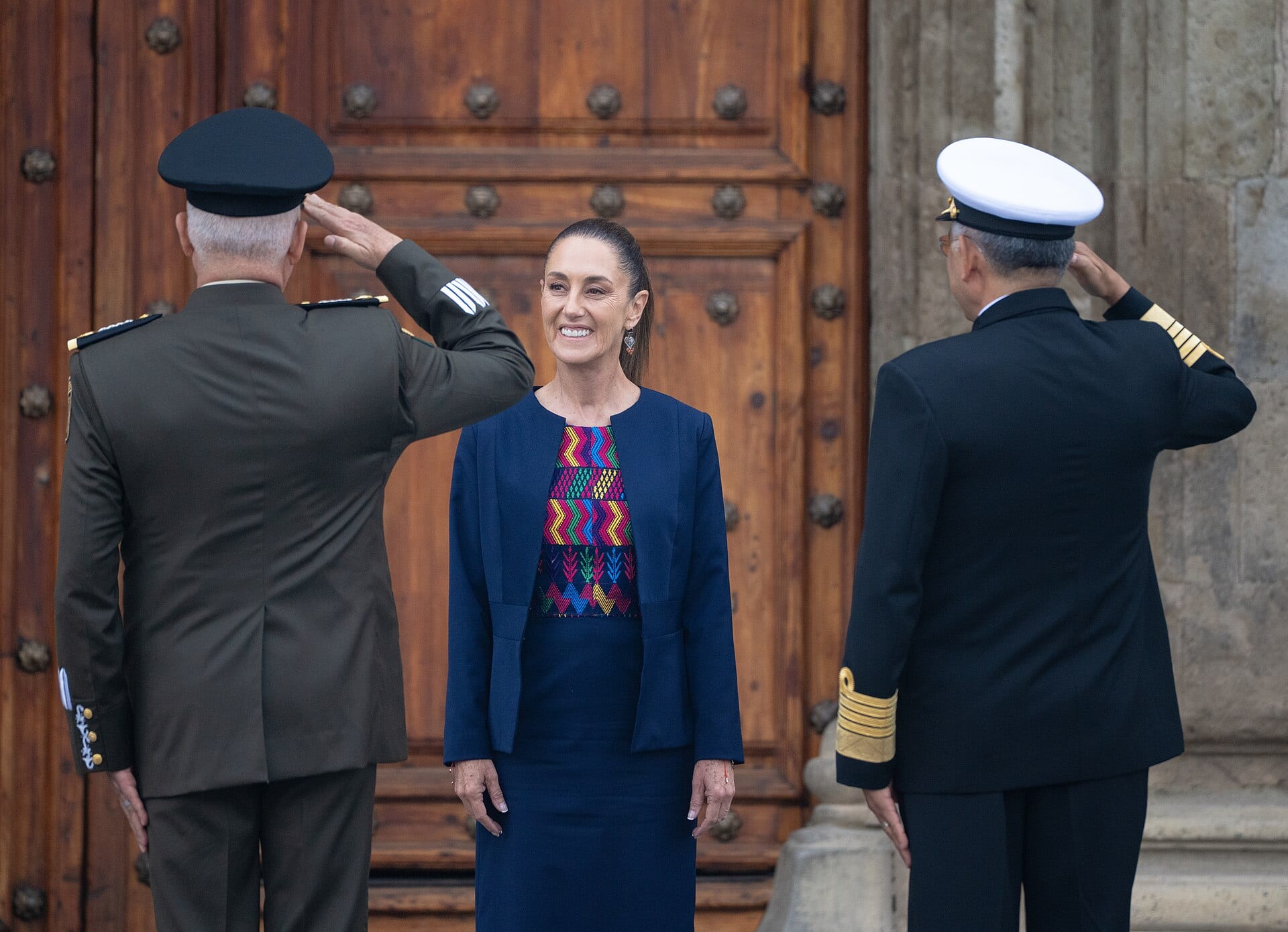 Claudia Sheinbaum sonríe de frente entre un militar del Ejército y un almirante de la Marina, ambos de espaldas y haciendo el saludo militar.