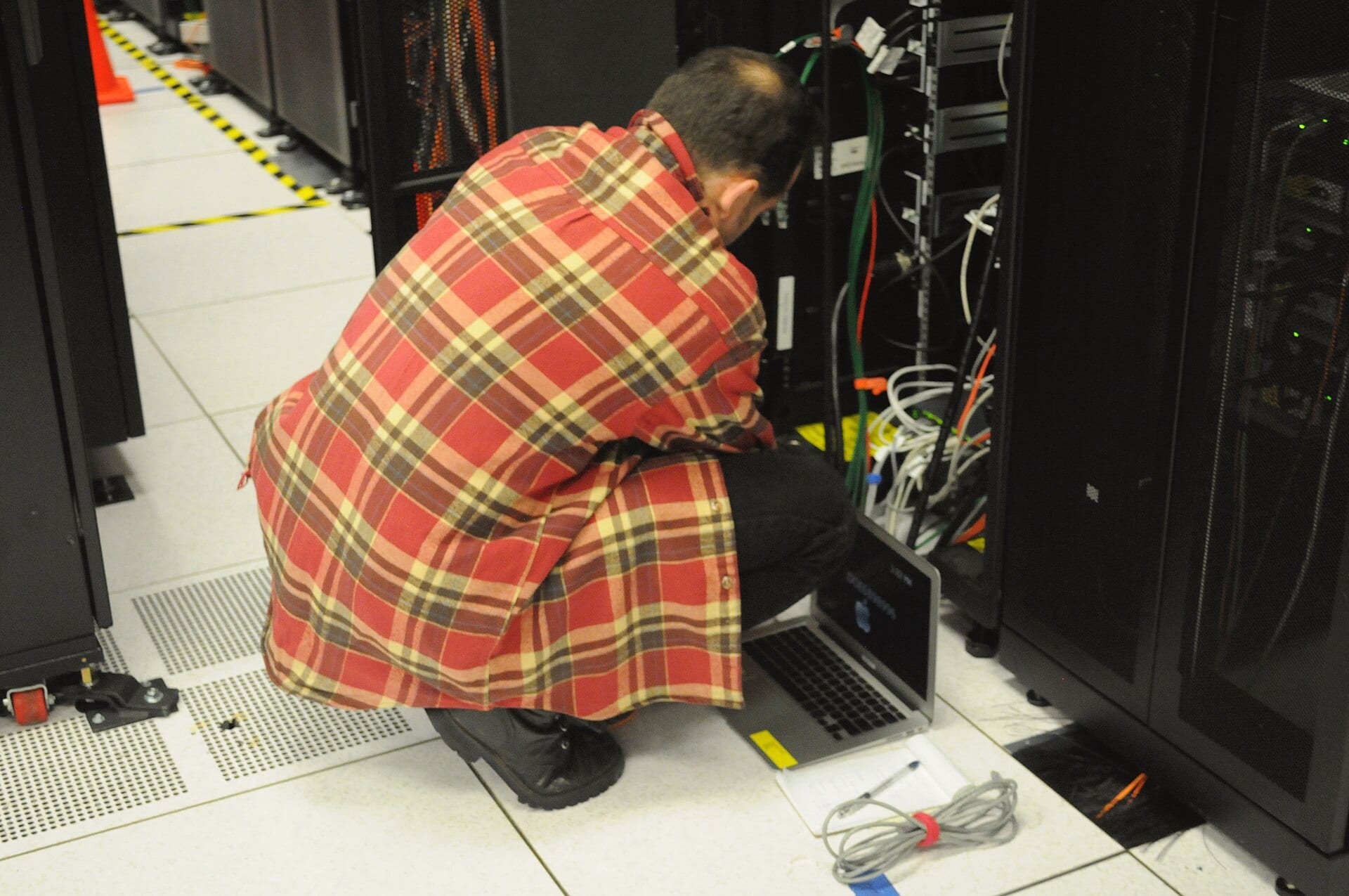 Un técnico en cuclillas conecta cables en la parte trasera de un rack de servidores, con una laptop abierta en el piso.