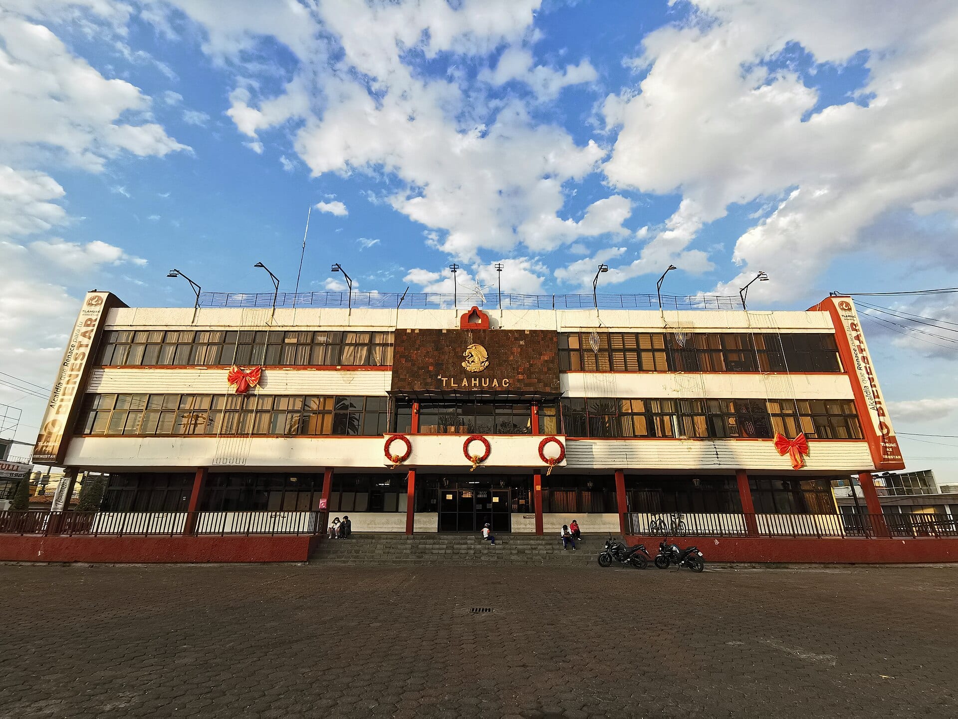 Fachada de dos pisos del edificio de la alcaldía de Tláhuac, blanca y roja, con el escudo nacional y el nombre de la demarcación sobre la entrada, bajo un cielo con nubes.