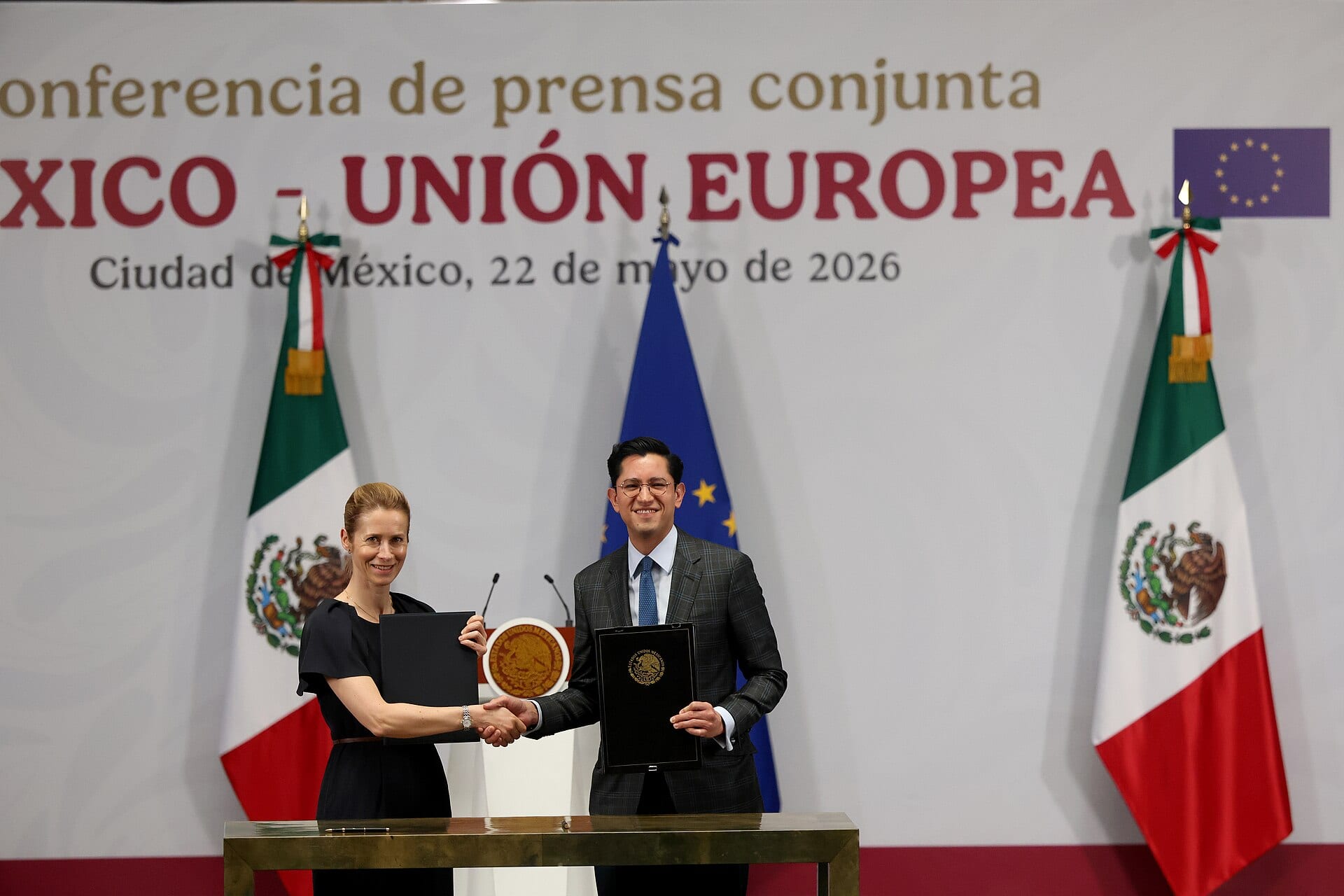 Una mujer de vestido oscuro y un hombre de traje gris con corbata azul se dan la mano ante banderas de México y de la Unión Europea.