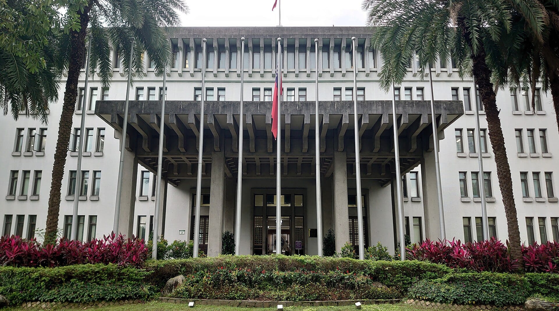 Fachada blanca de un edificio de cuatro pisos con un gran porche de concreto, palmeras a los lados y una bandera roja ondeando frente a la entrada.