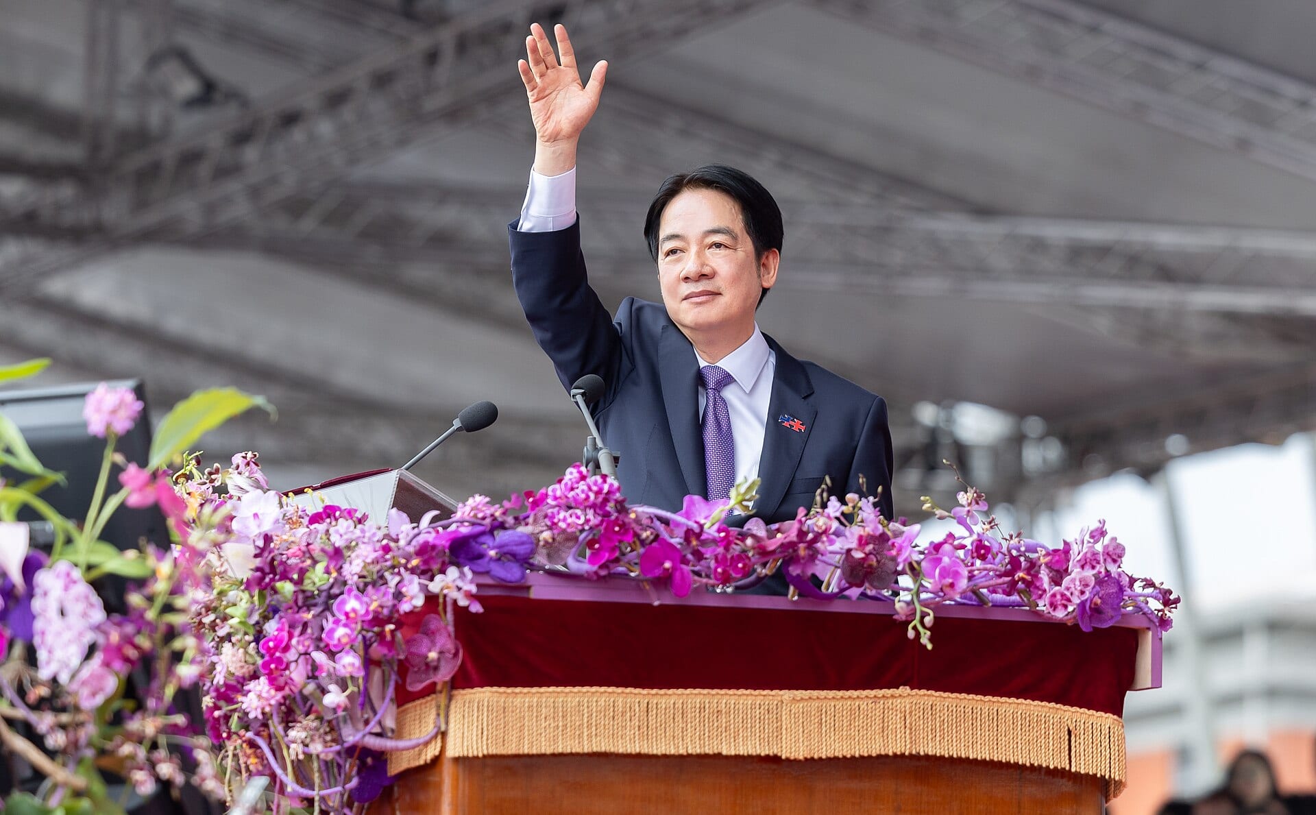 Lai Ching-te, in a dark suit, waves with a raised hand behind a podium covered in purple and pink orchids.